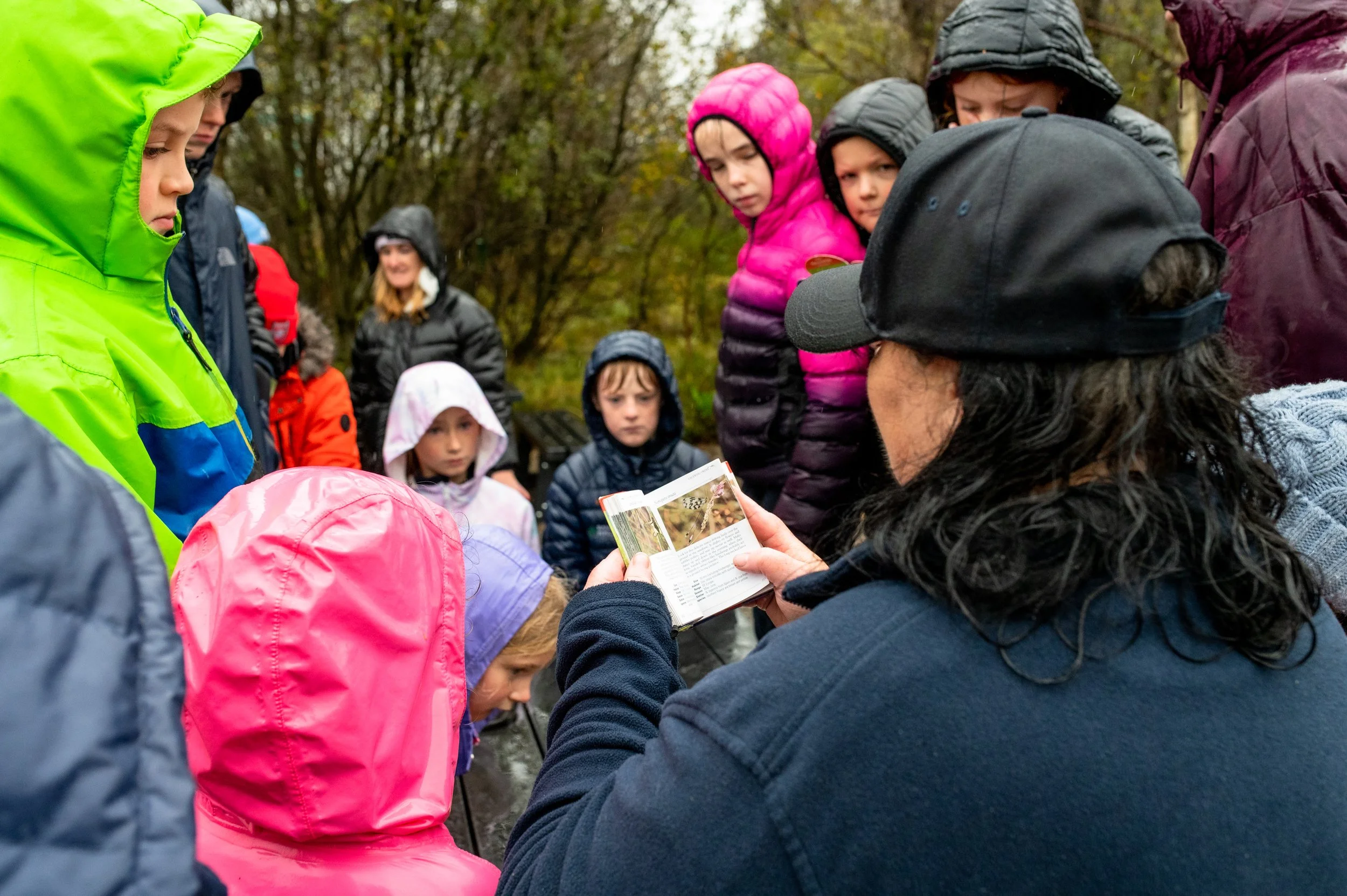 Group of children and an adult outdoors, wearing rain jackets and hoods, engaged in an educational activity, with trees in the background.