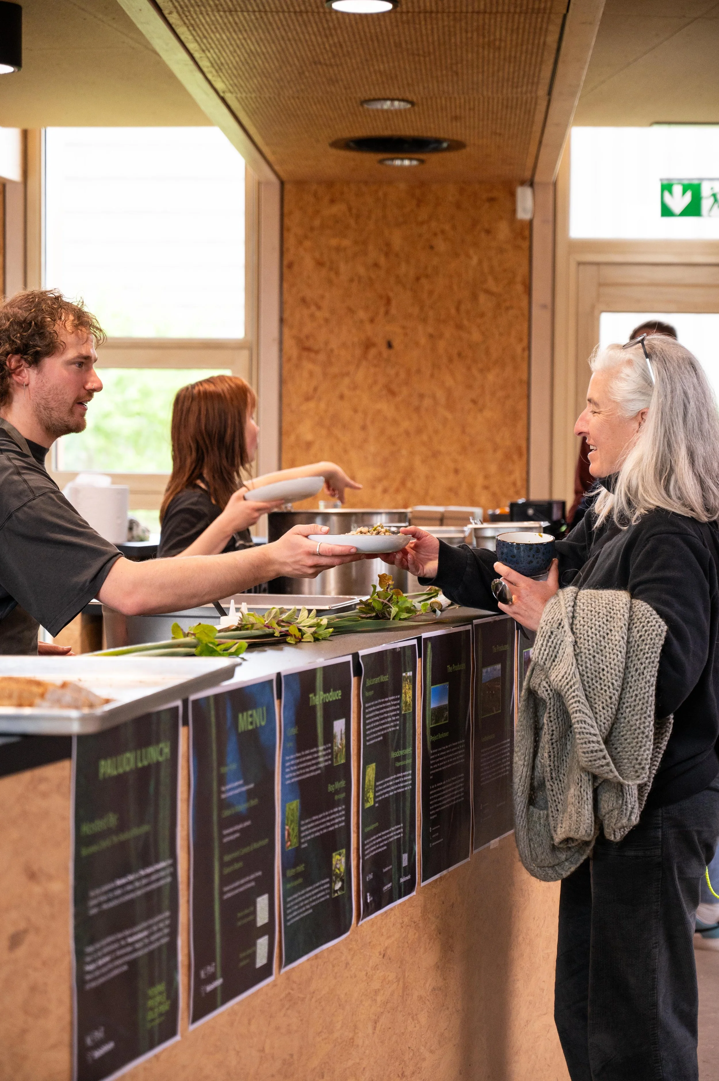 A woman with gray hair wearing glasses and a gray sweater receiving a bowl of food from a man with curly brown hair, both standing in front of a display of informational posters on a wall at an indoor event.