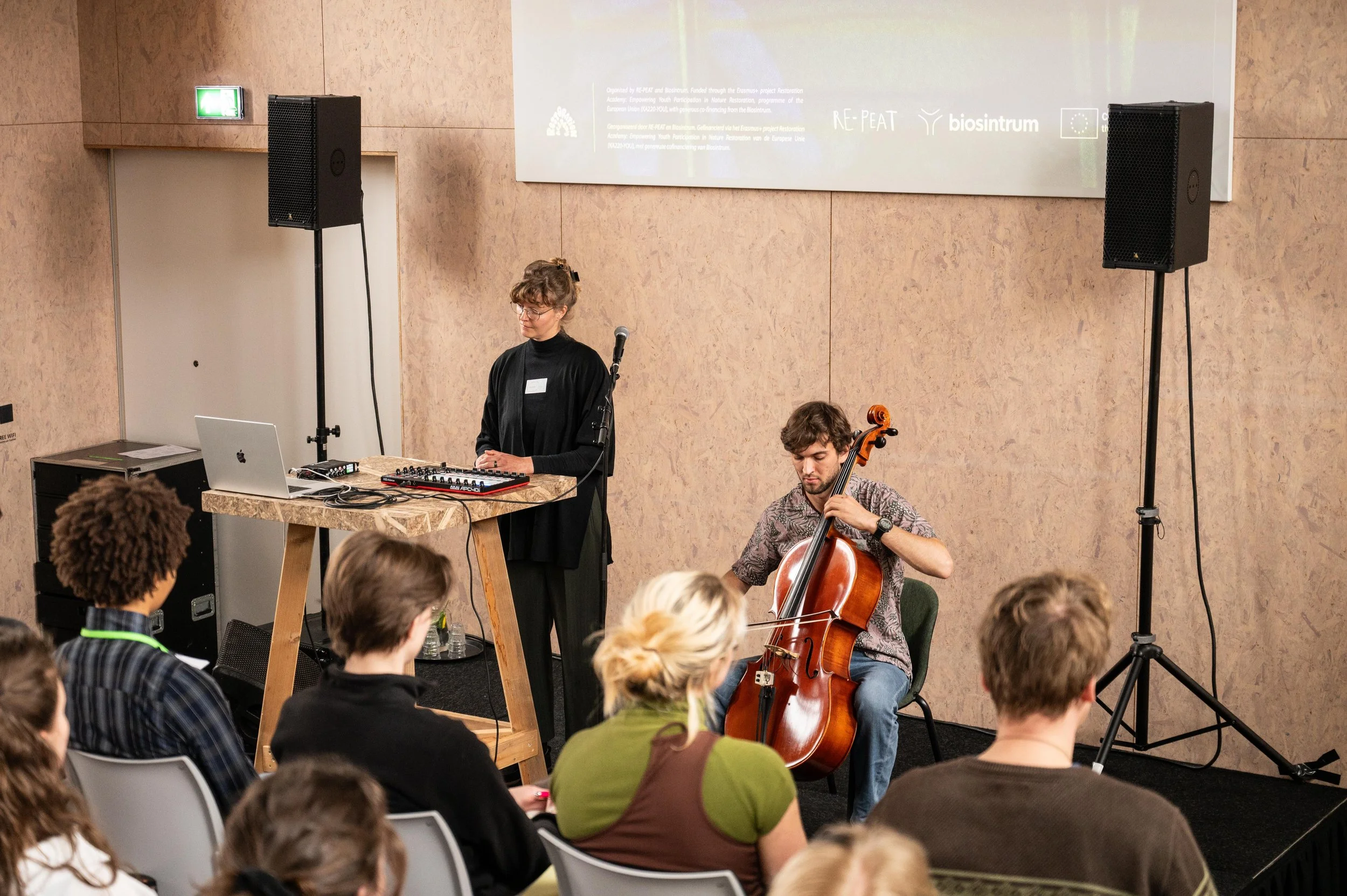 Musicians performing at an indoor event with an audience. A woman stands at a DJ setup with a laptop and mixer, while a man plays a cello beside her. The audience watches attentively.