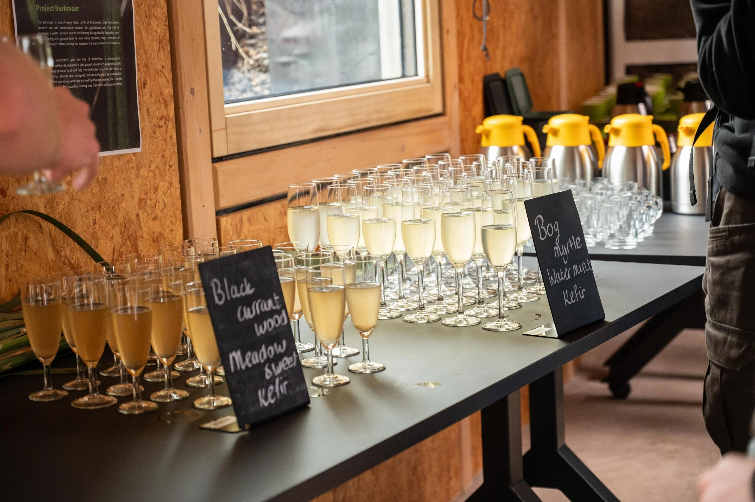 A display of multiple glasses of champagne on a table, with chalkboard signs indicating flavors like 'Black currant' and 'Water mint,' in a rustic indoor setting with wood-paneled walls.
