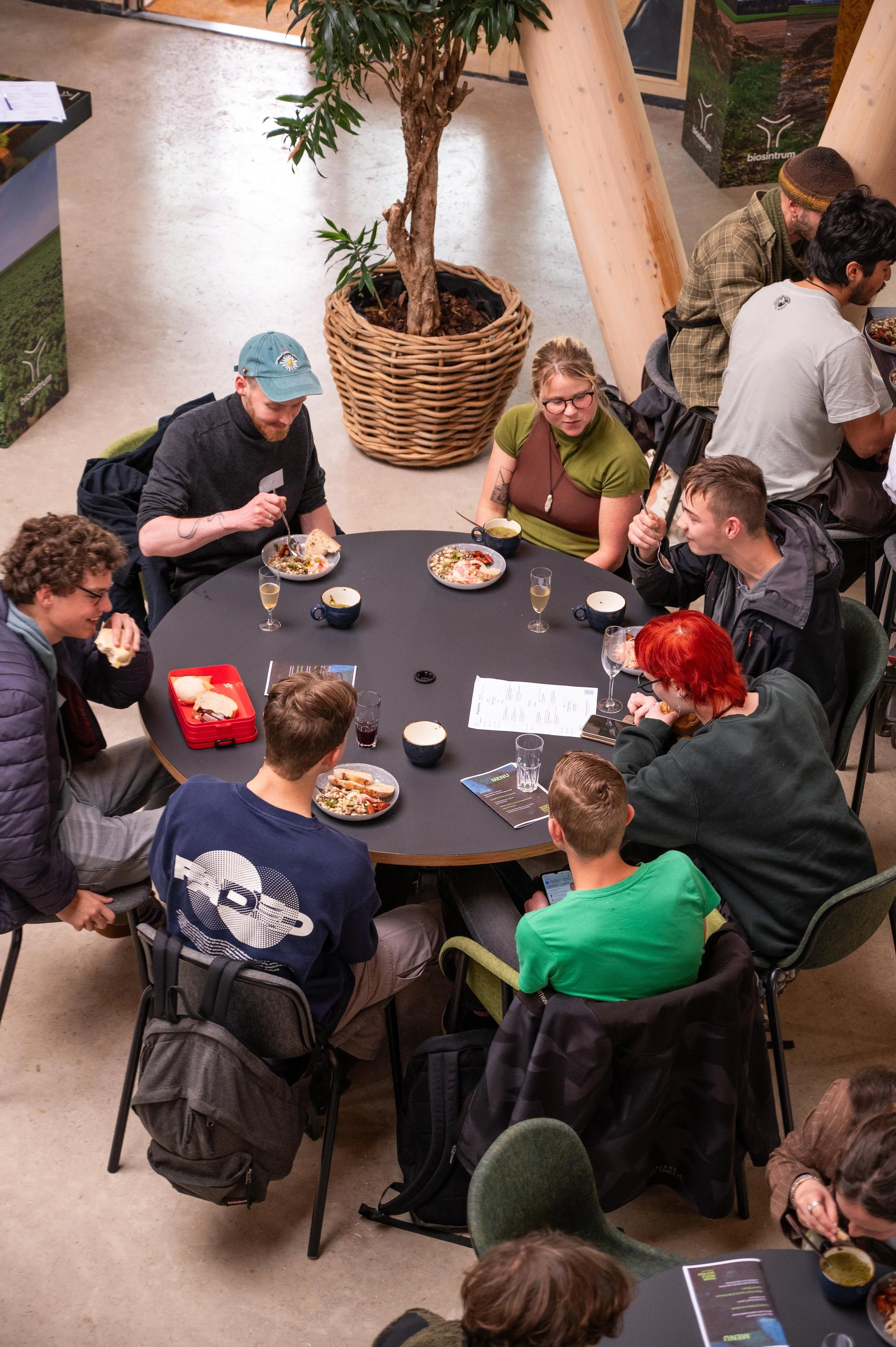 People sitting around a black round table enjoying a meal in a room with wooden support beams and potted plant, some with backpacks on chairs.