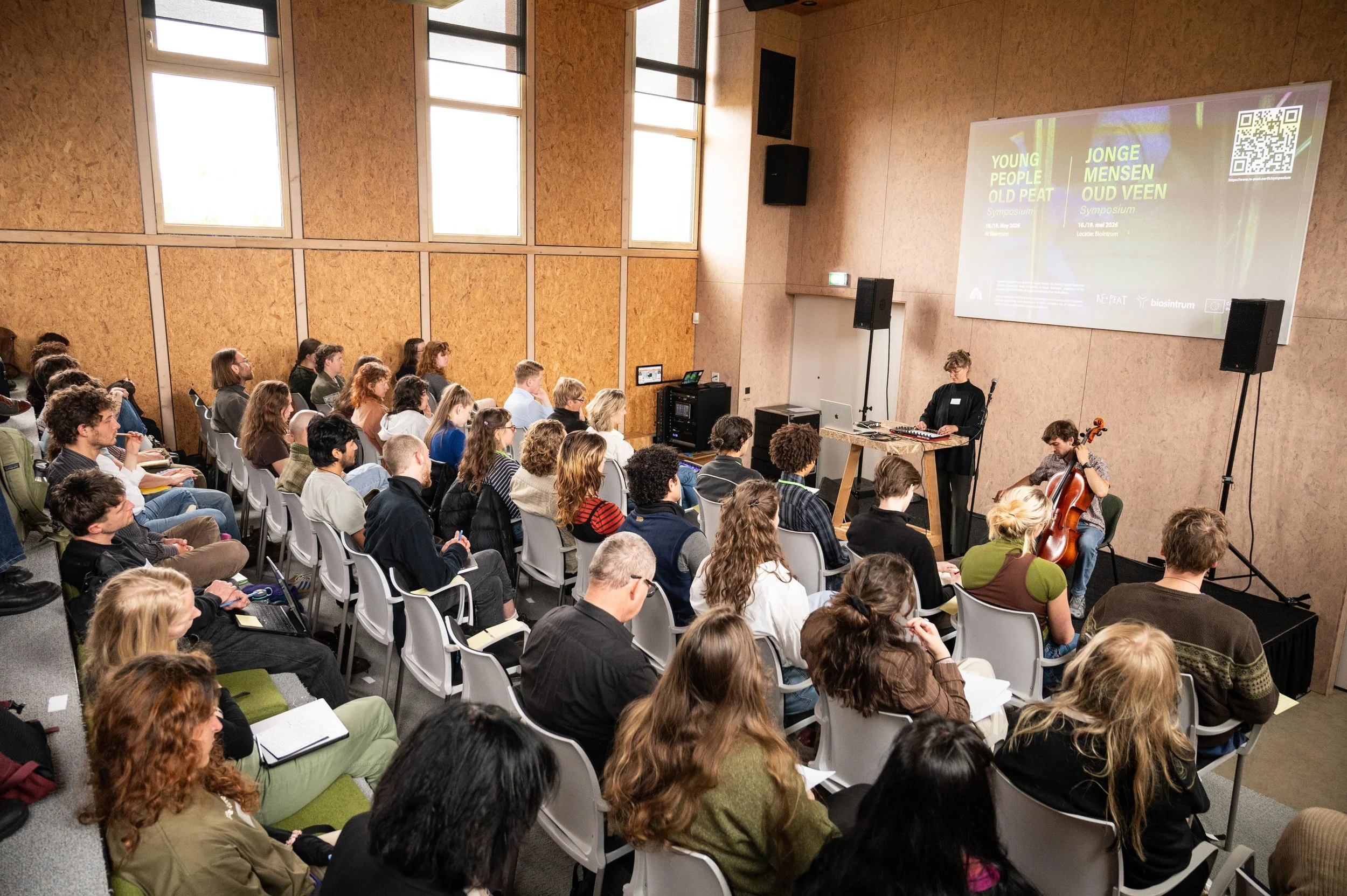 A large audience attending a symposium in a spacious room with high windows, where a presentation is displayed on a large screen at the front. The event is focused on youth and old peat, and features a musician playing cello.