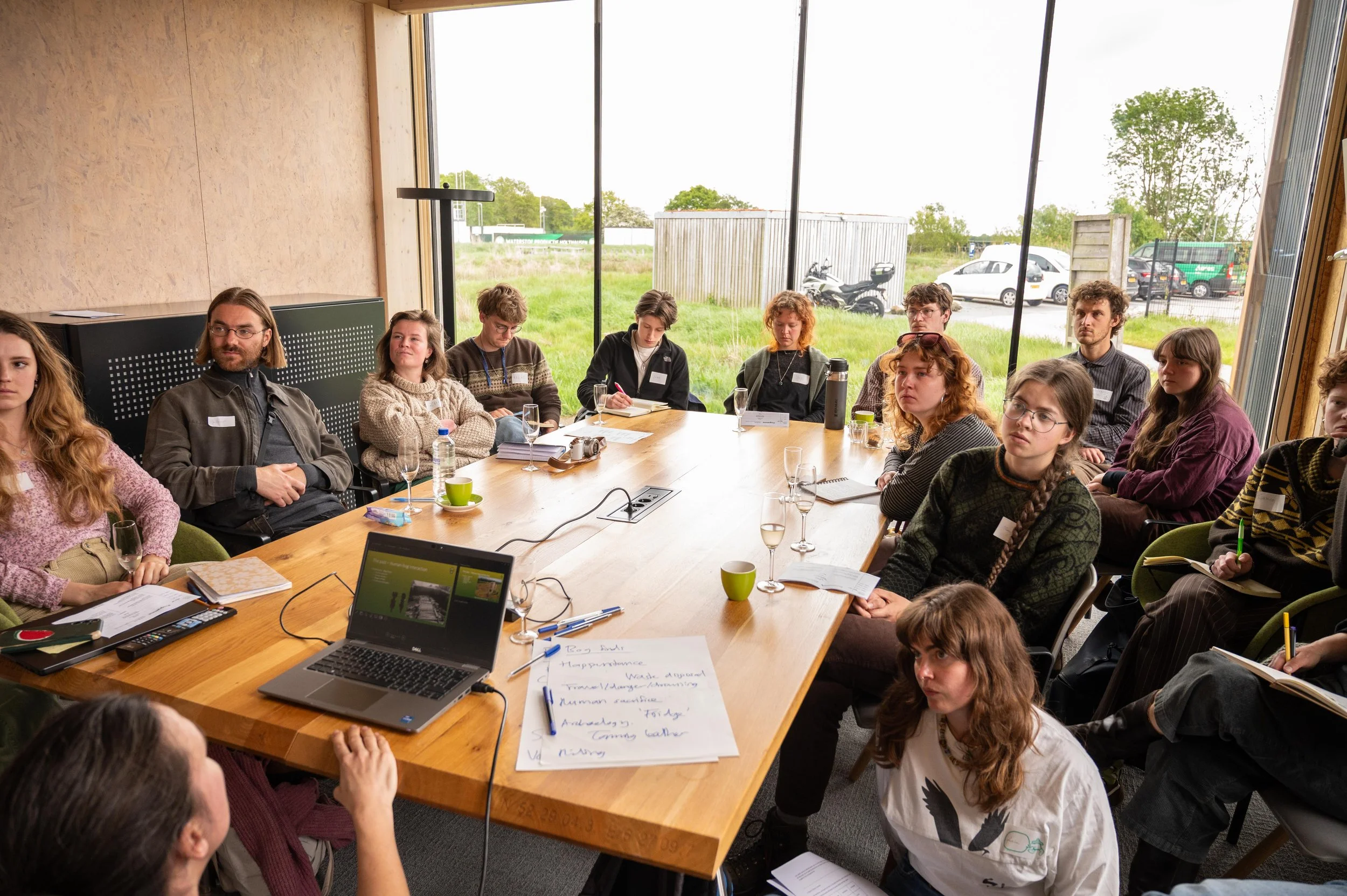 A group of people sitting around a large wooden table in a modern conference room with floor-to-ceiling windows showing a grassy outside area and parked cars. The table has laptops, notebooks, glasses, and other conference materials.