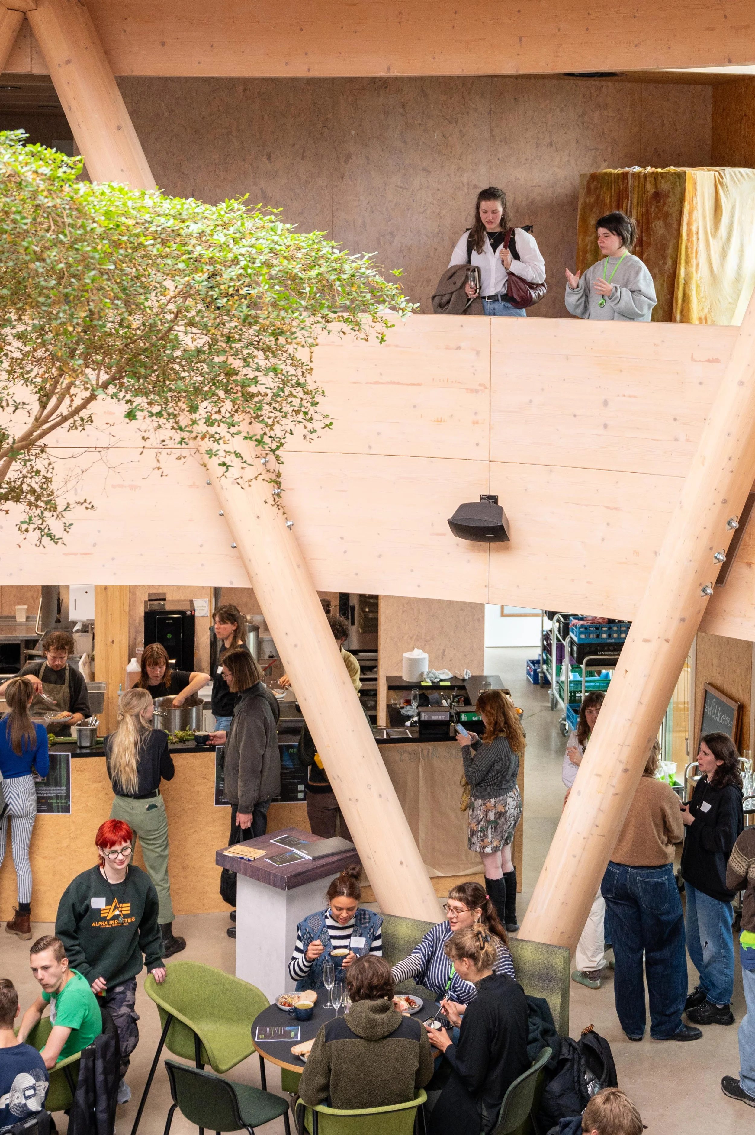 People gathered in a busy indoor restaurant or cafe, some standing and chatting, others seated and eating. The setting features wooden beams, a tree, and a second-story balcony or platform.