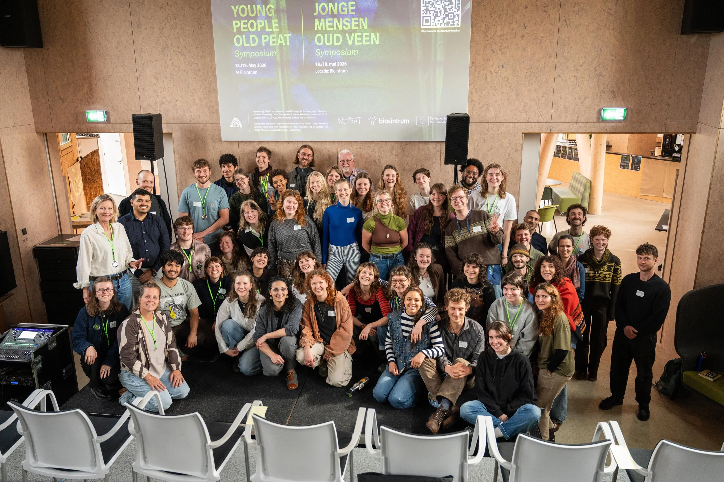 Group photo of diverse young people and adults gathered in a large indoor space, smiling and posing for the camera at a symposium event about young and old peats, held on May 18-19, 2026, with a large presentation screen behind them.