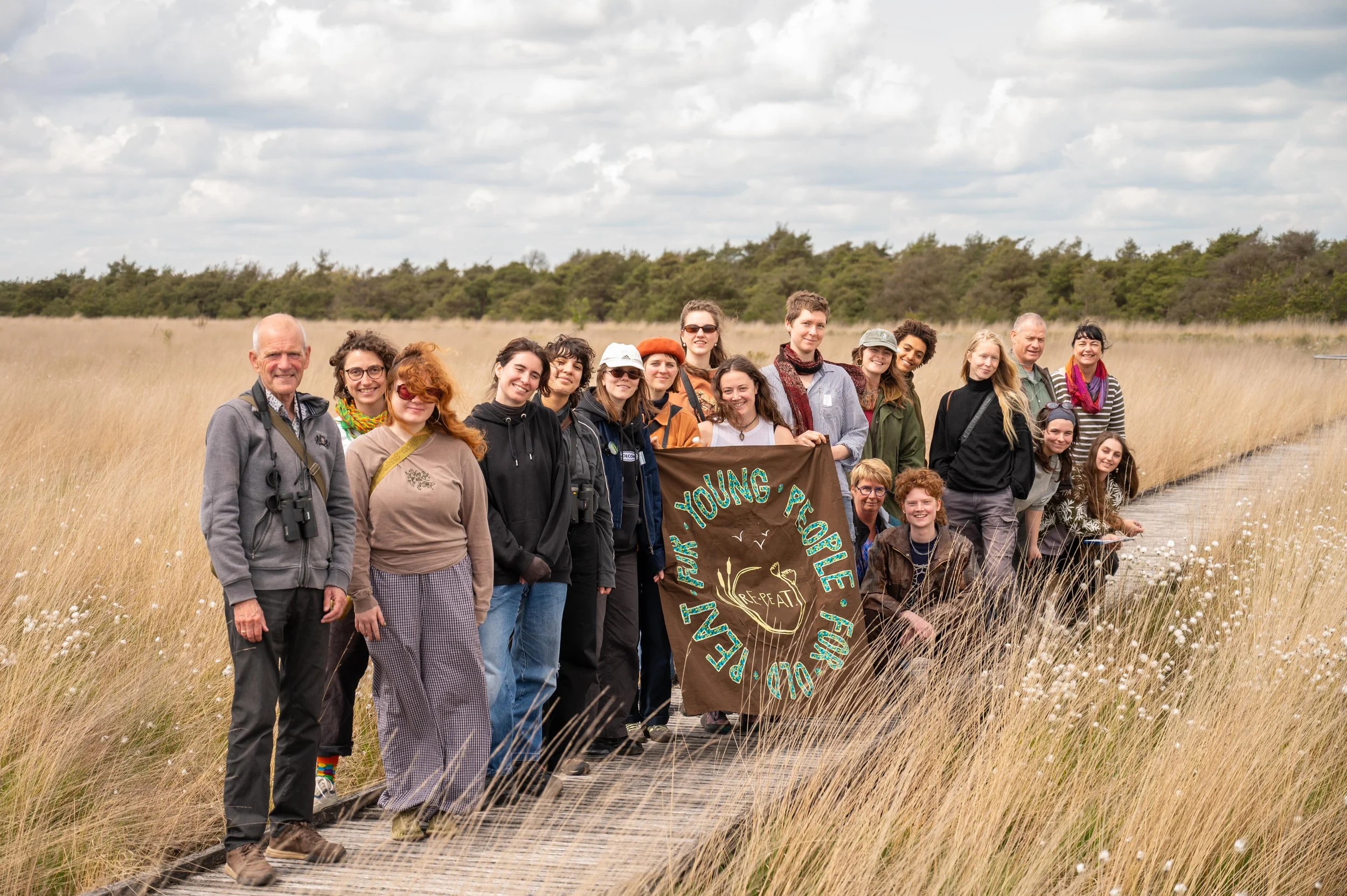 Group of people standing outdoors in a grassy field during cloudy weather, holding a brown banner with green and yellow text that reads 'Young People' and other words, possibly part of a natural or environmental event.