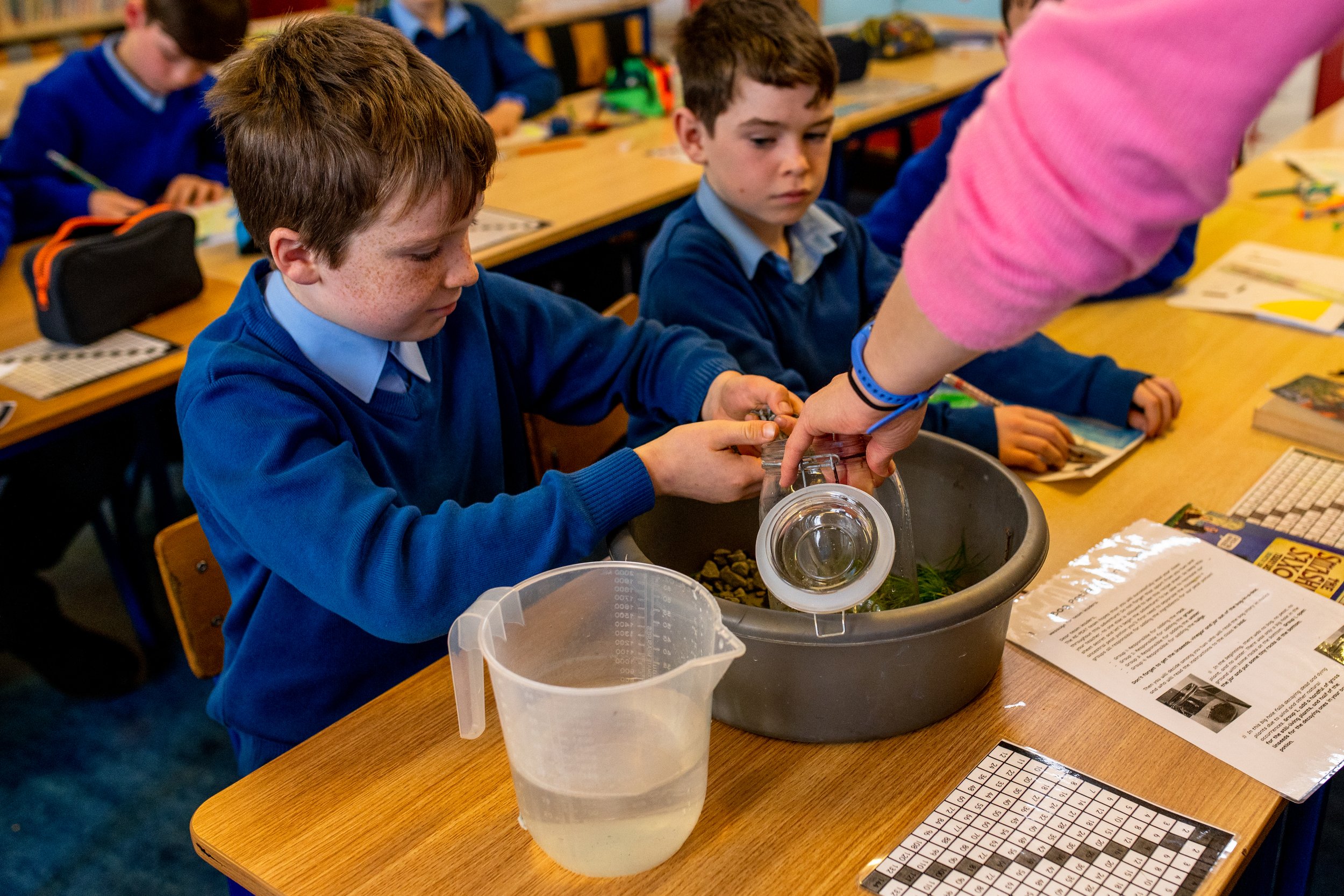 Students in classroom conducting a science experiment with a teacher, mixing substances in a large bowl, surrounded by educational materials.