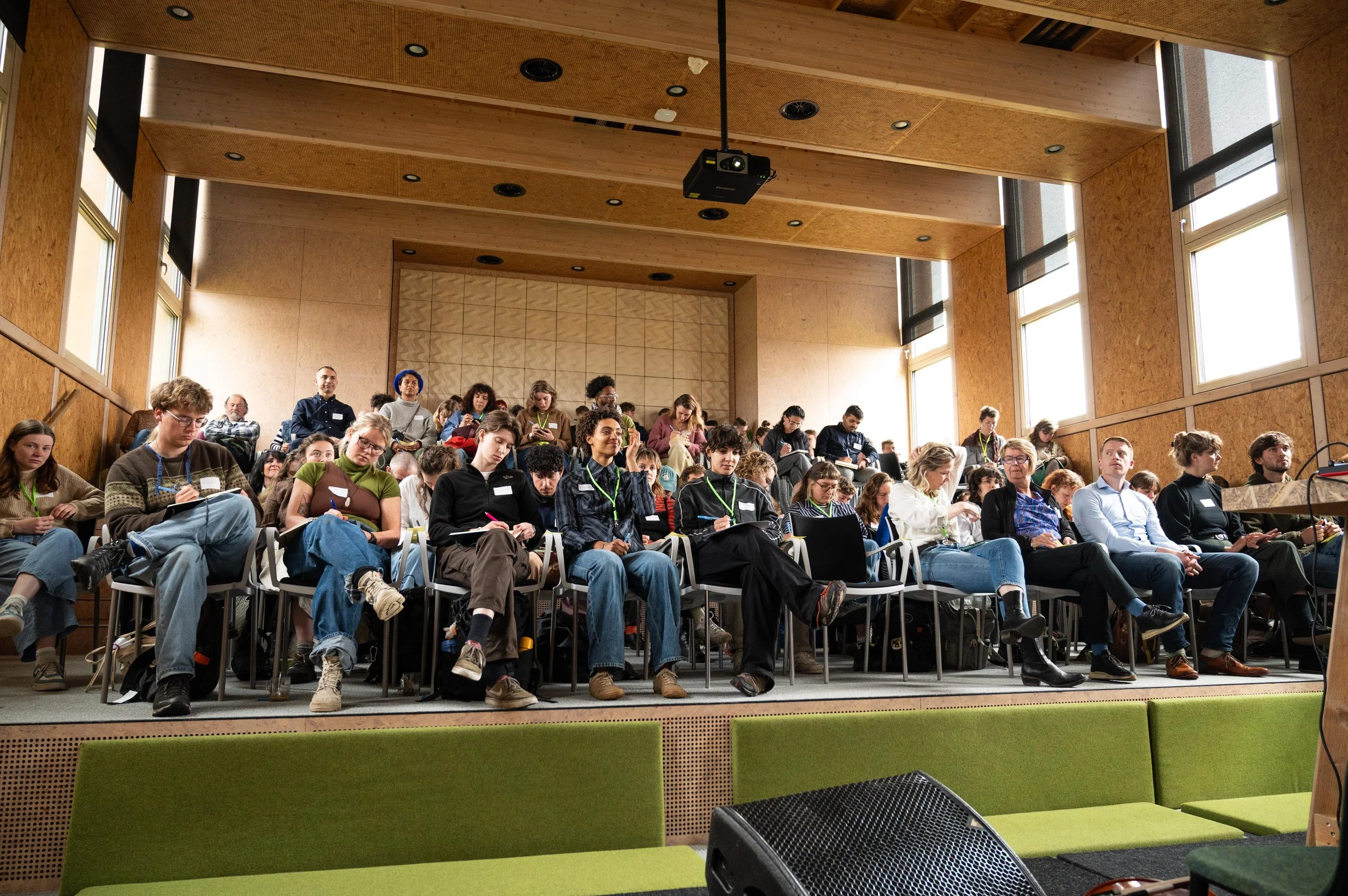 Audience seated in a conference hall, taking notes, facing a stage with a green couch and a speaker at a podium.