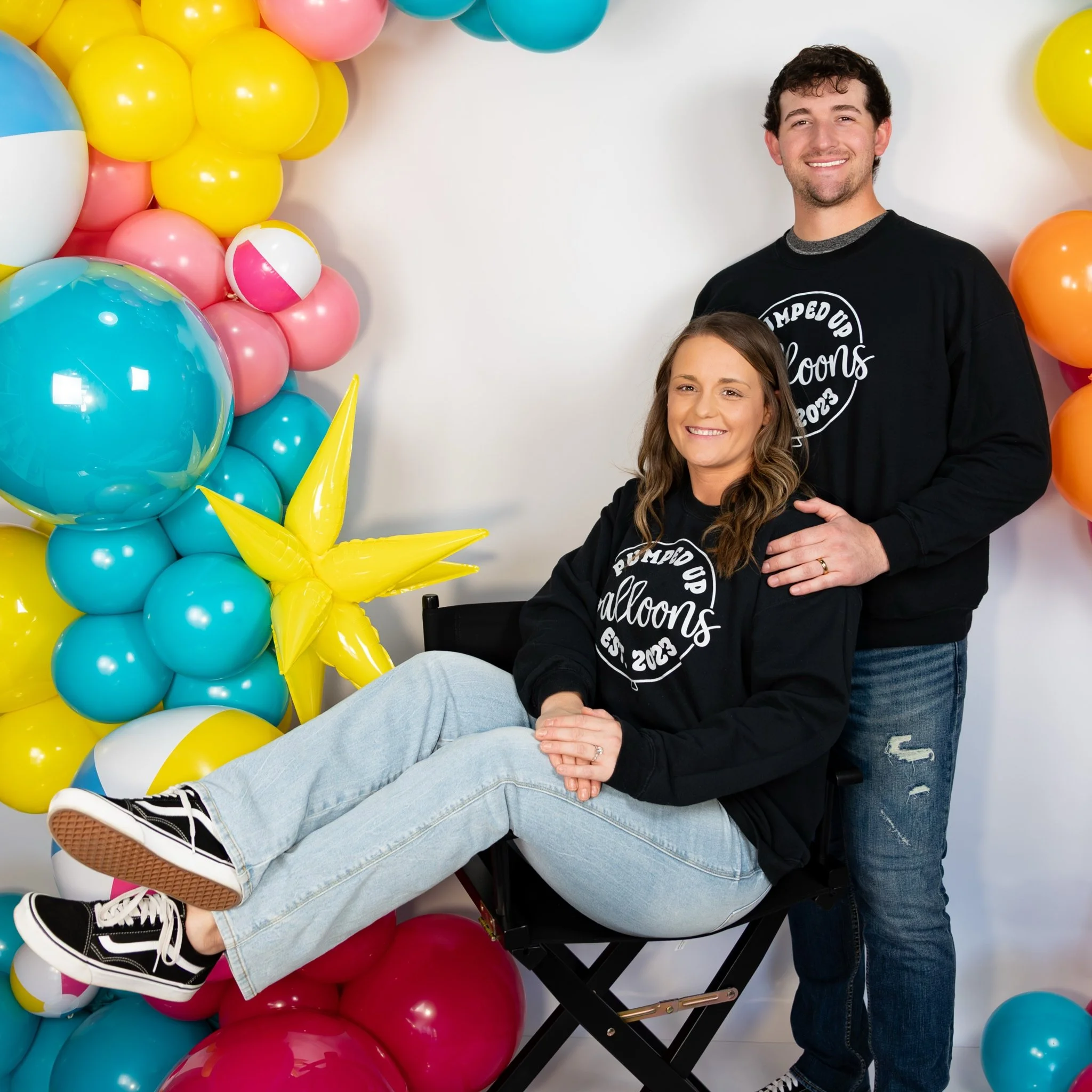 A man standing behind a woman sitting in a director's chair, both smiling, surrounded by colorful balloons in shades of yellow, pink, blue, orange, and white, at a balloon sculpting event.