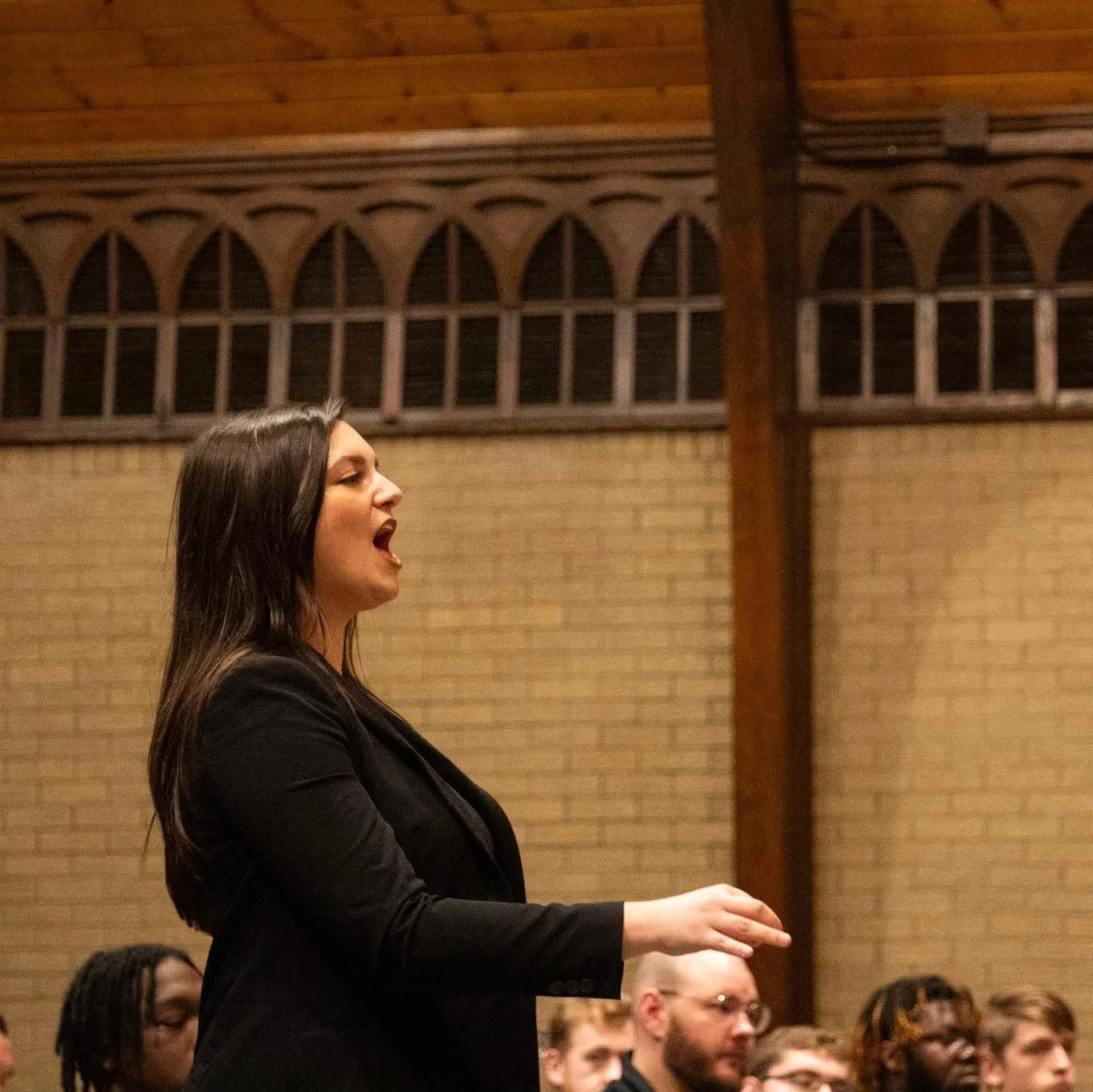 A woman with long dark hair wearing a black jacket speaking passionately to an audience in a room with brick walls and wooden ceiling.