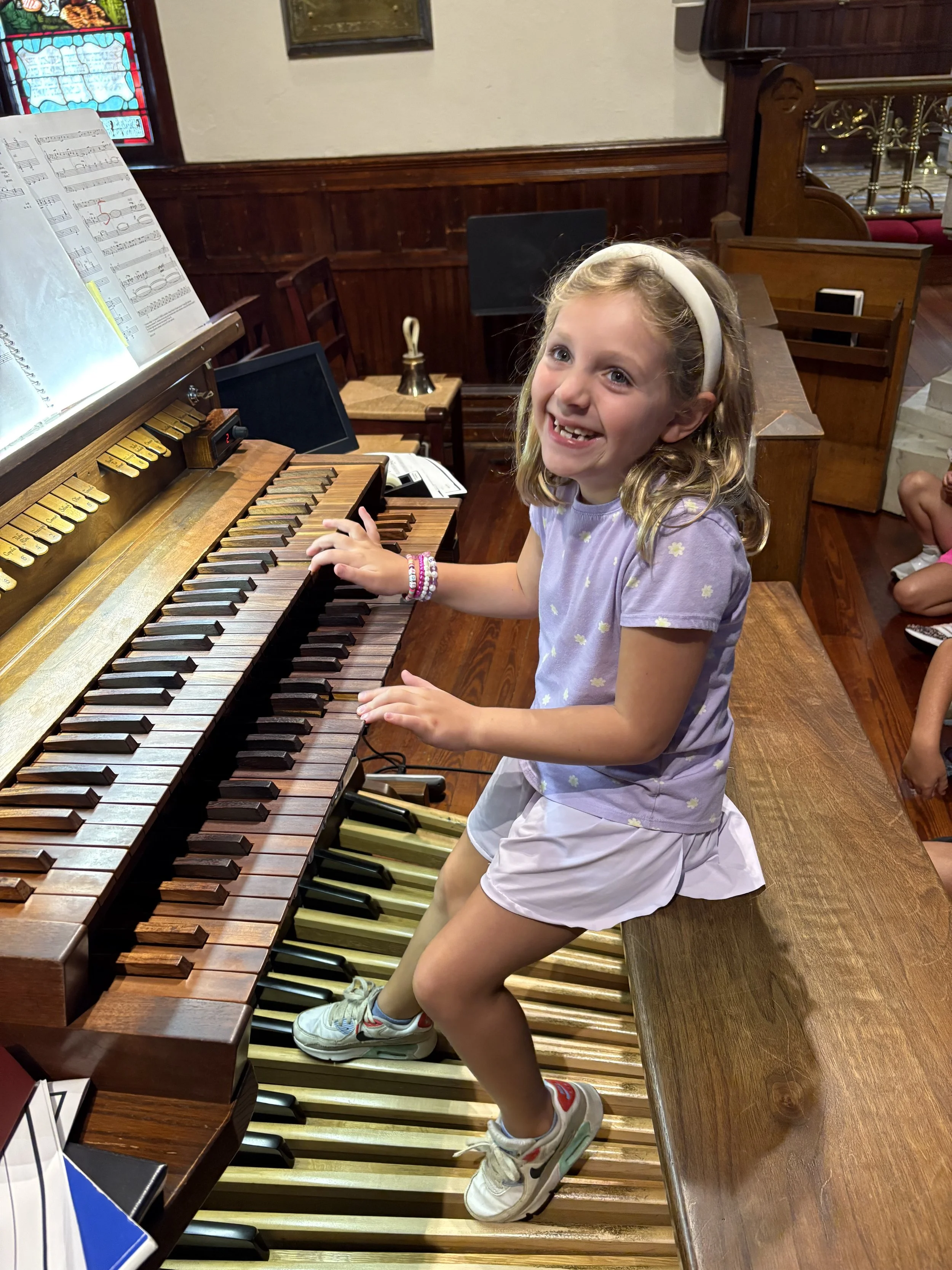 A young girl with blonde hair, wearing a lavender shirt with a daisy pattern, white shorts, and sneakers, sitting at a wooden piano in a church. She has a big smile and is touching the piano keys with her left hand, looking at the camera.
