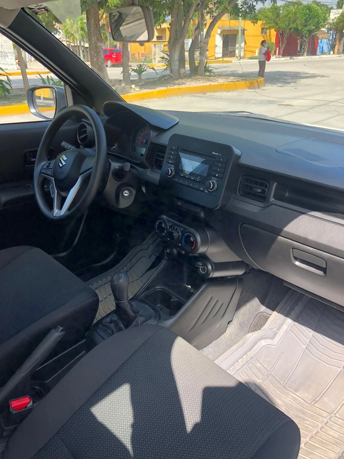 Interior of a Suzuki car showing the steering wheel, dashboard, gear shift, and front seats. Outside view through the windshield shows a street with trees, a woman on the sidewalk, and colorful buildings.