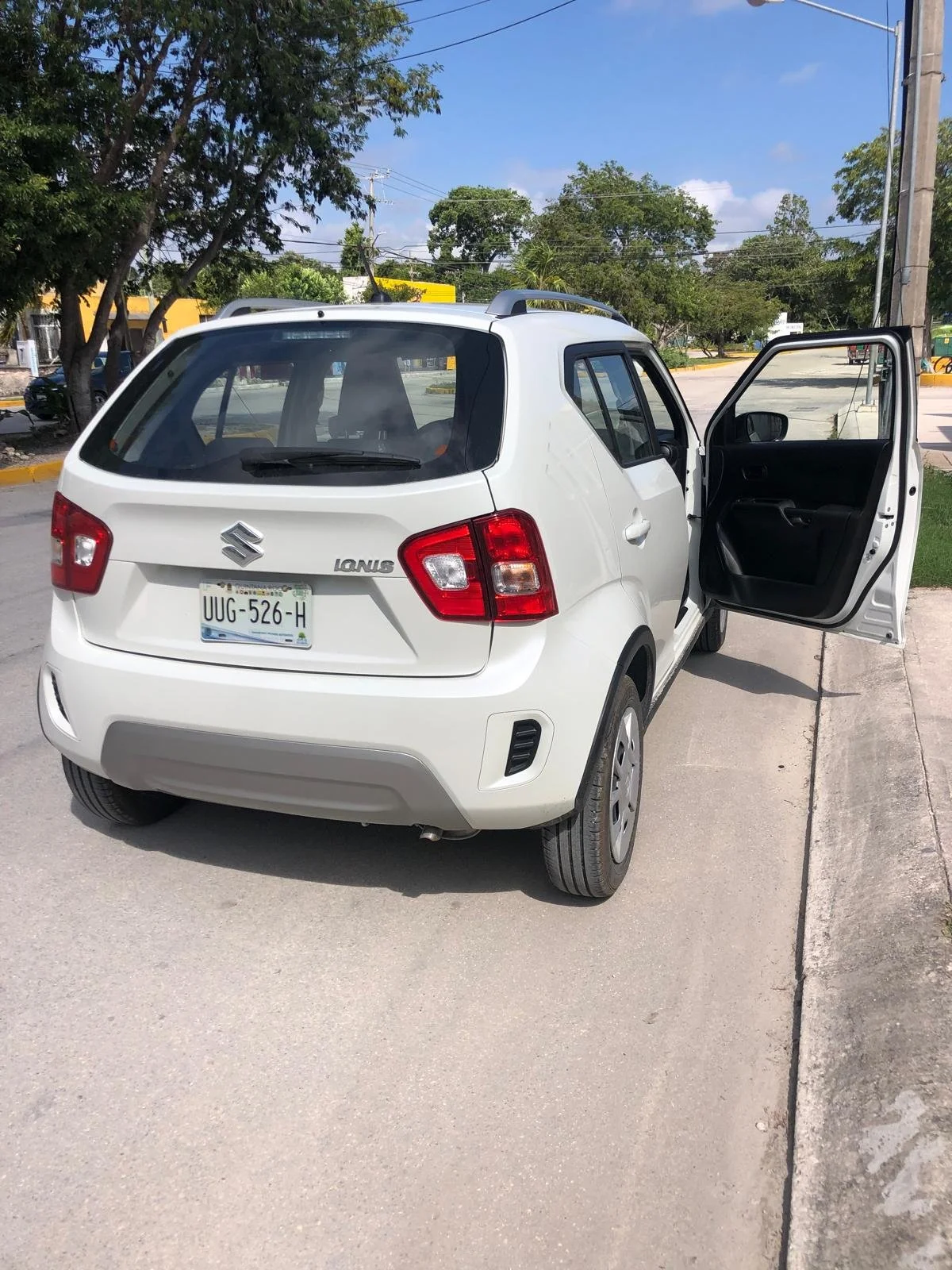 White Suzuki Ignis parked on the side of a street with its driver's door open, sunny weather, trees and a clear blue sky in the background.