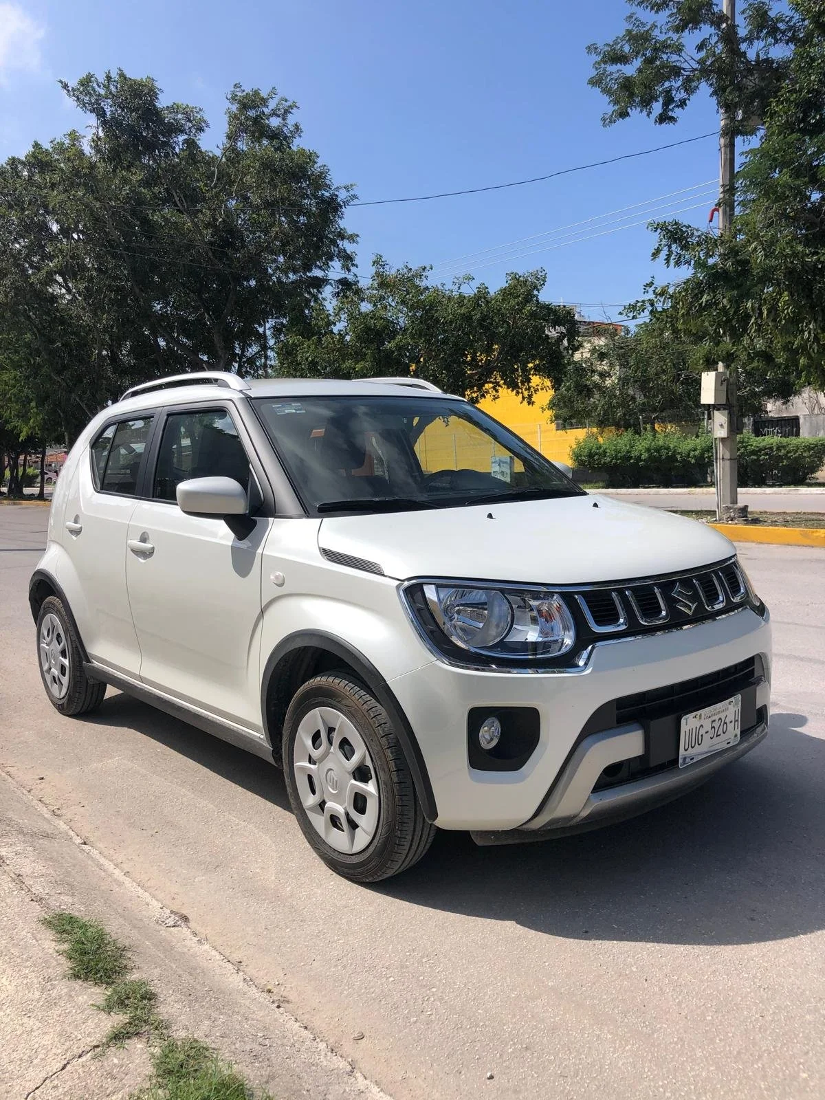 A white compact SUV parked on a street near trees and a yellow building under a clear blue sky.