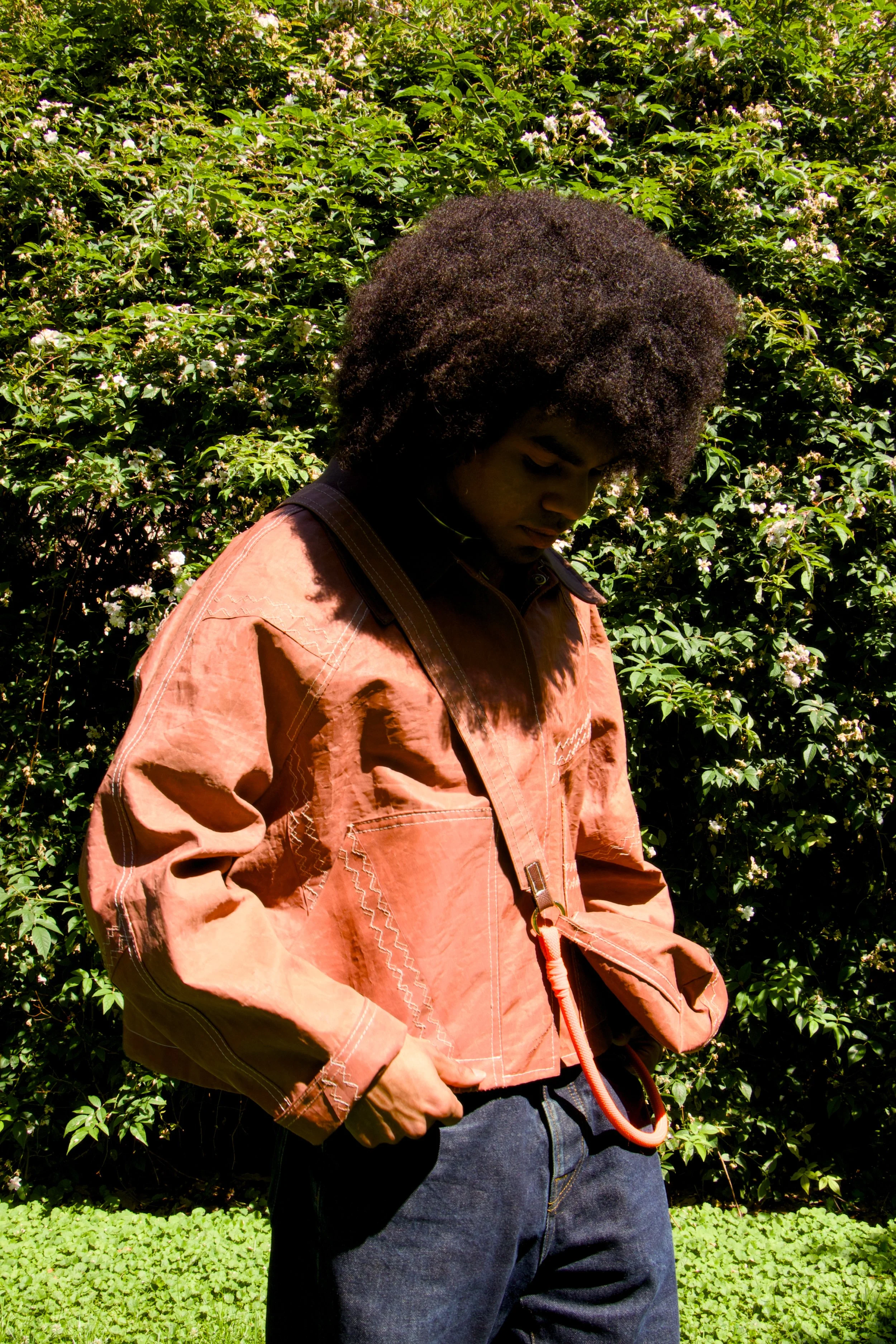 A person with dark skin and curly afro hair looking down, wearing a dusty rose jacket with a strap and jeans, standing outdoors in front of a green bush with small white flowers.