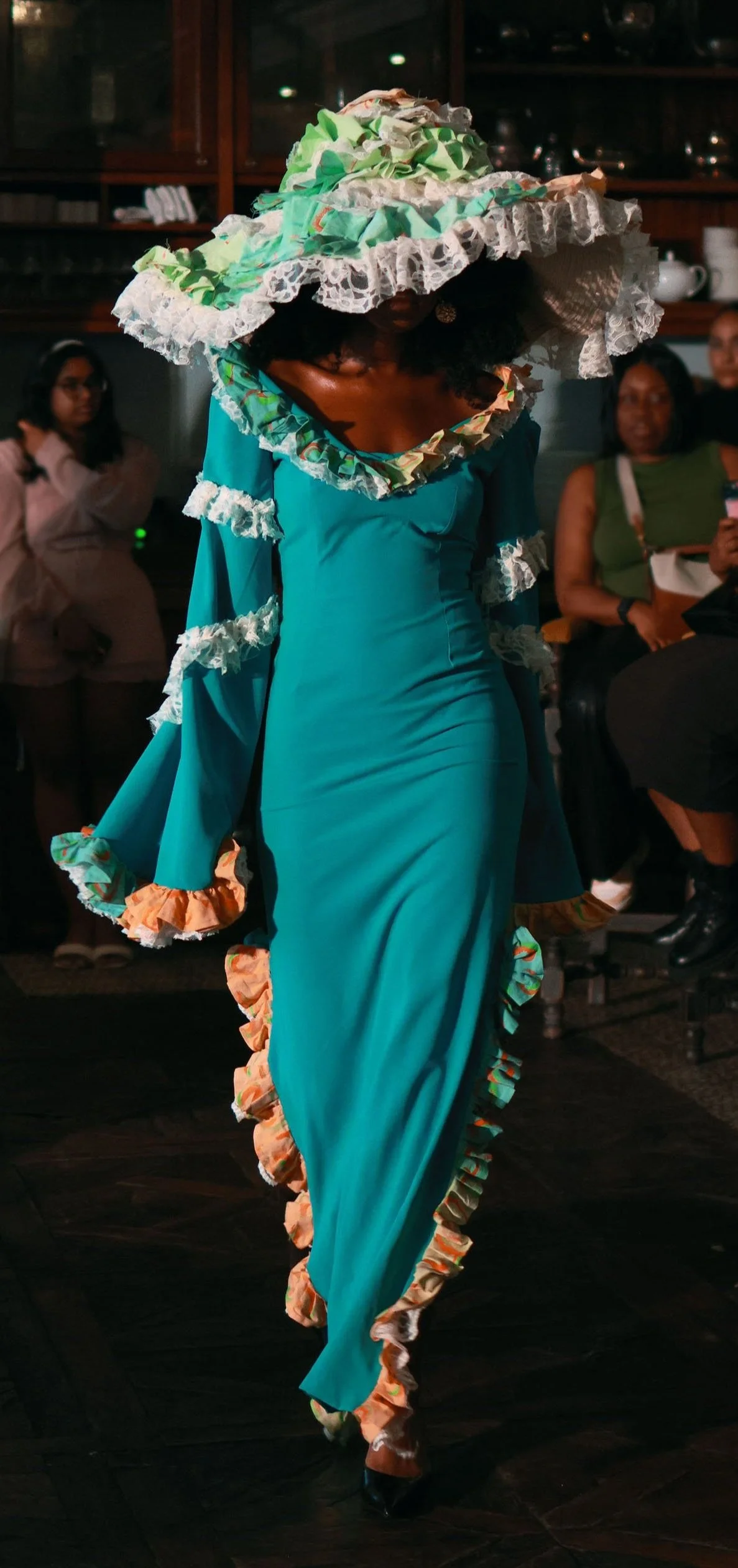 A woman wearing a teal dress with ruffles and a large, elaborate hat is walking on a dark wooden floor in front of seated and standing people in a dimly lit room.