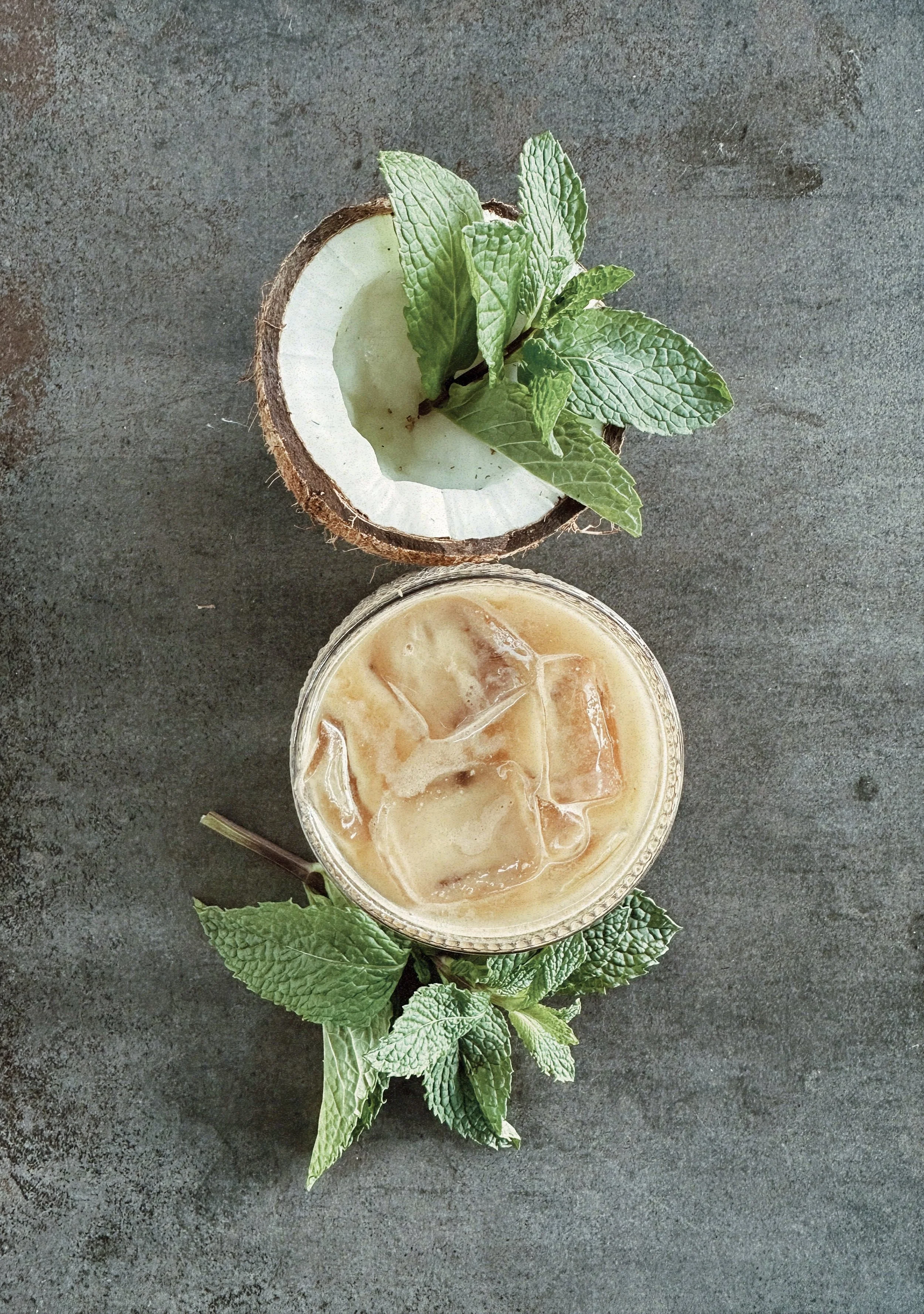 Top view of a coconut half filled with mint leaves and ice, next to a glass of iced coffee with ice and mint leaves on a dark textured surface.