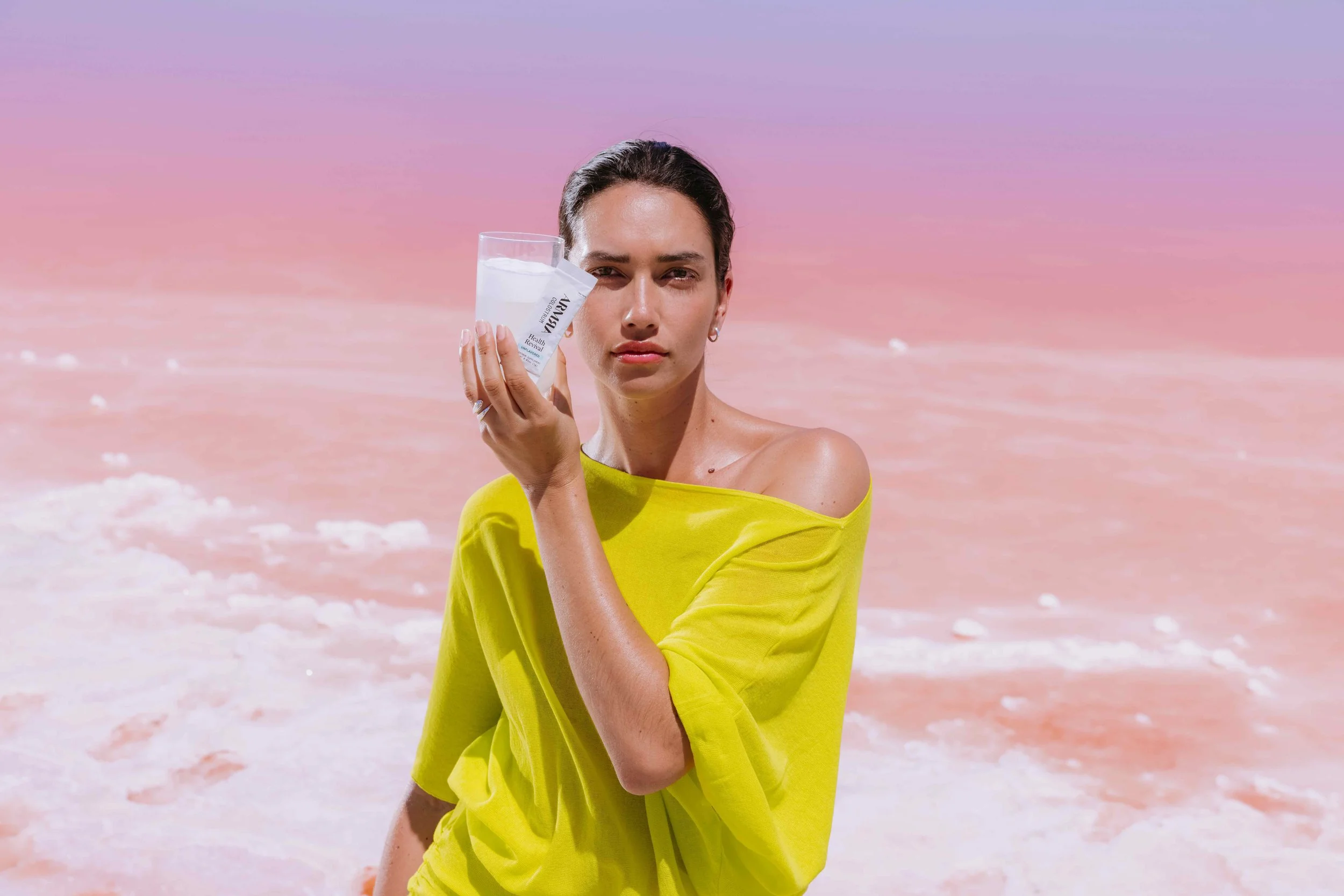 Woman with dark hair in a yellow off-the-shoulder top holding a glass of water against a pink and purple sky at the beach.