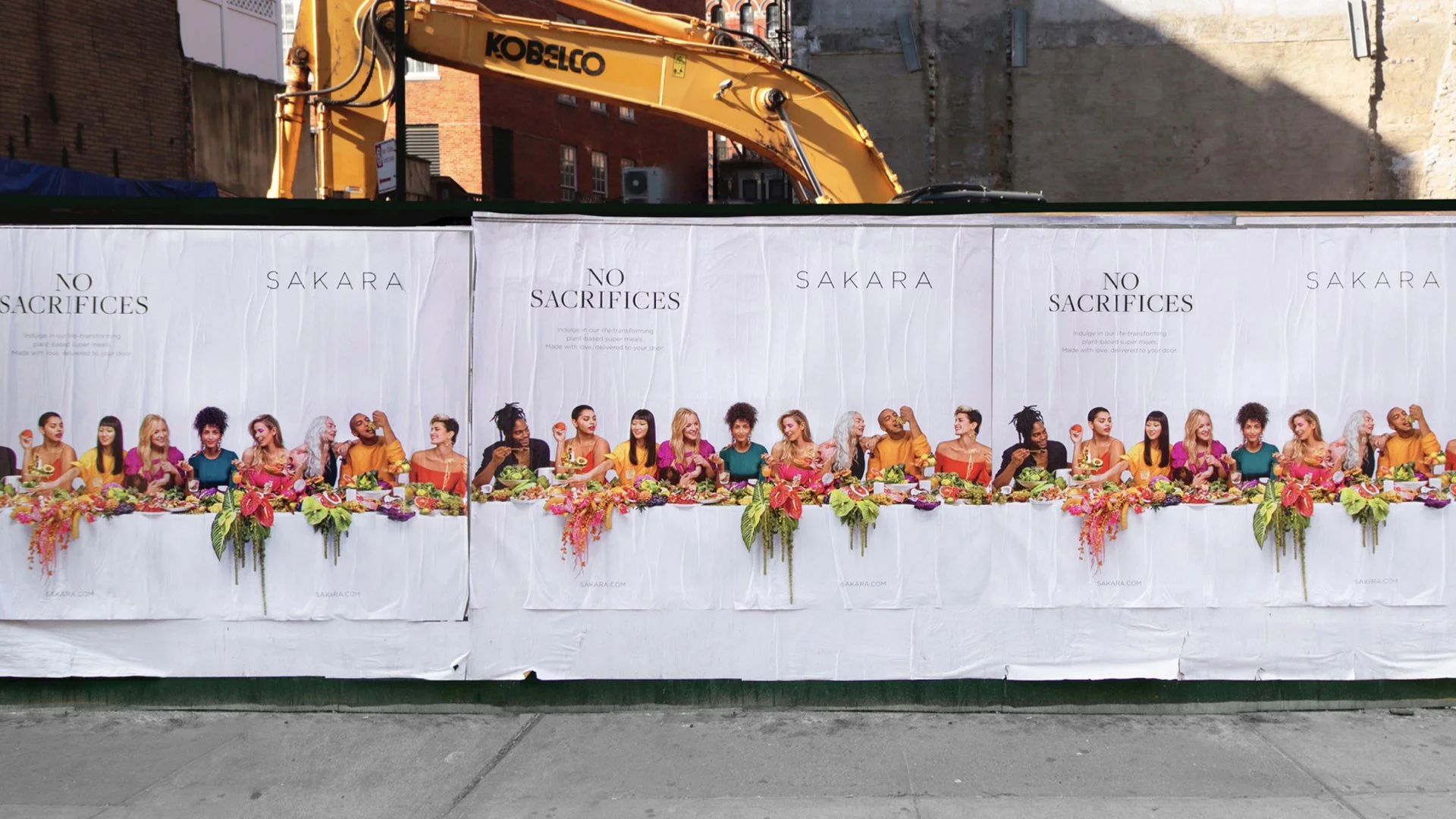 A long table with colorful flowers and various food dishes, set against white banners with text, with diverse women seated and engaged as if at a communal meal or celebration.