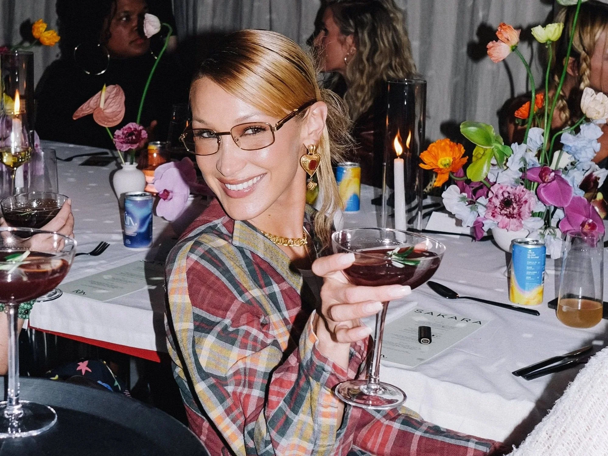 A woman with light skin, wearing glasses, gold heart-shaped earrings, and a plaid shirt, smiling and holding a wine glass at a dinner party with floral arrangements and other guests in the background. Bella Hadid for Kin 