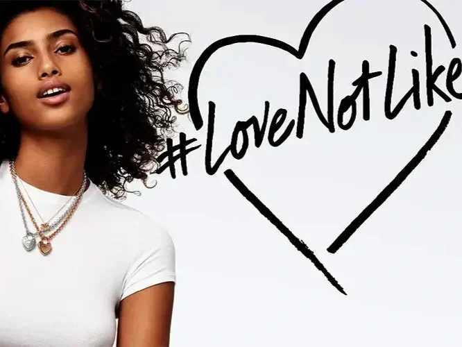 A woman with curly hair is standing next to a sign that reads '#LoveNotLike' inside a heart shape.