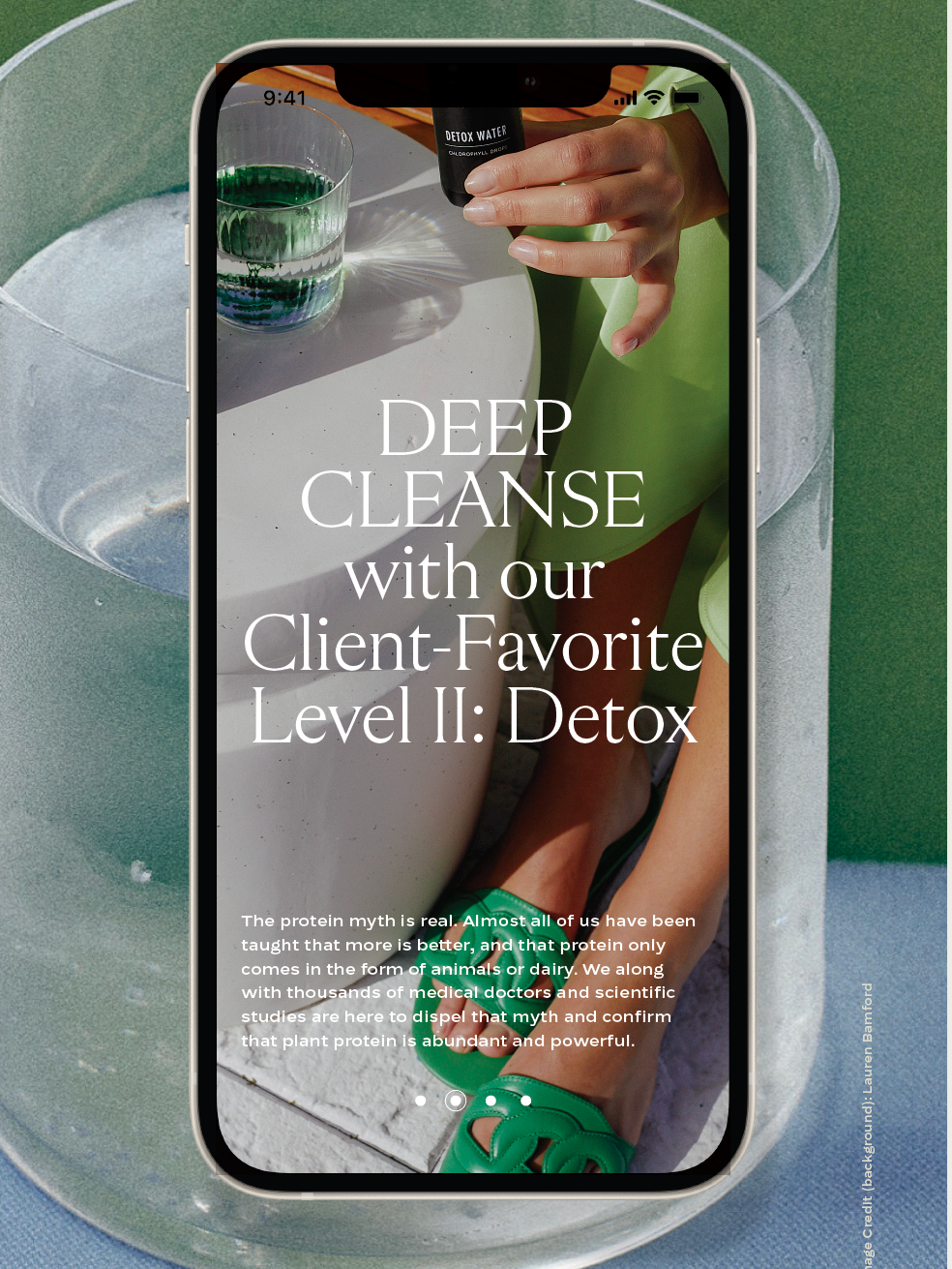 Smartphone displaying an advertisement for a detox water, with a glass of green liquid and a hand holding a small black bottle labeled 'Detox Water.' The phone is placed on a glass bowl, with a pool or outdoor setting in the background.