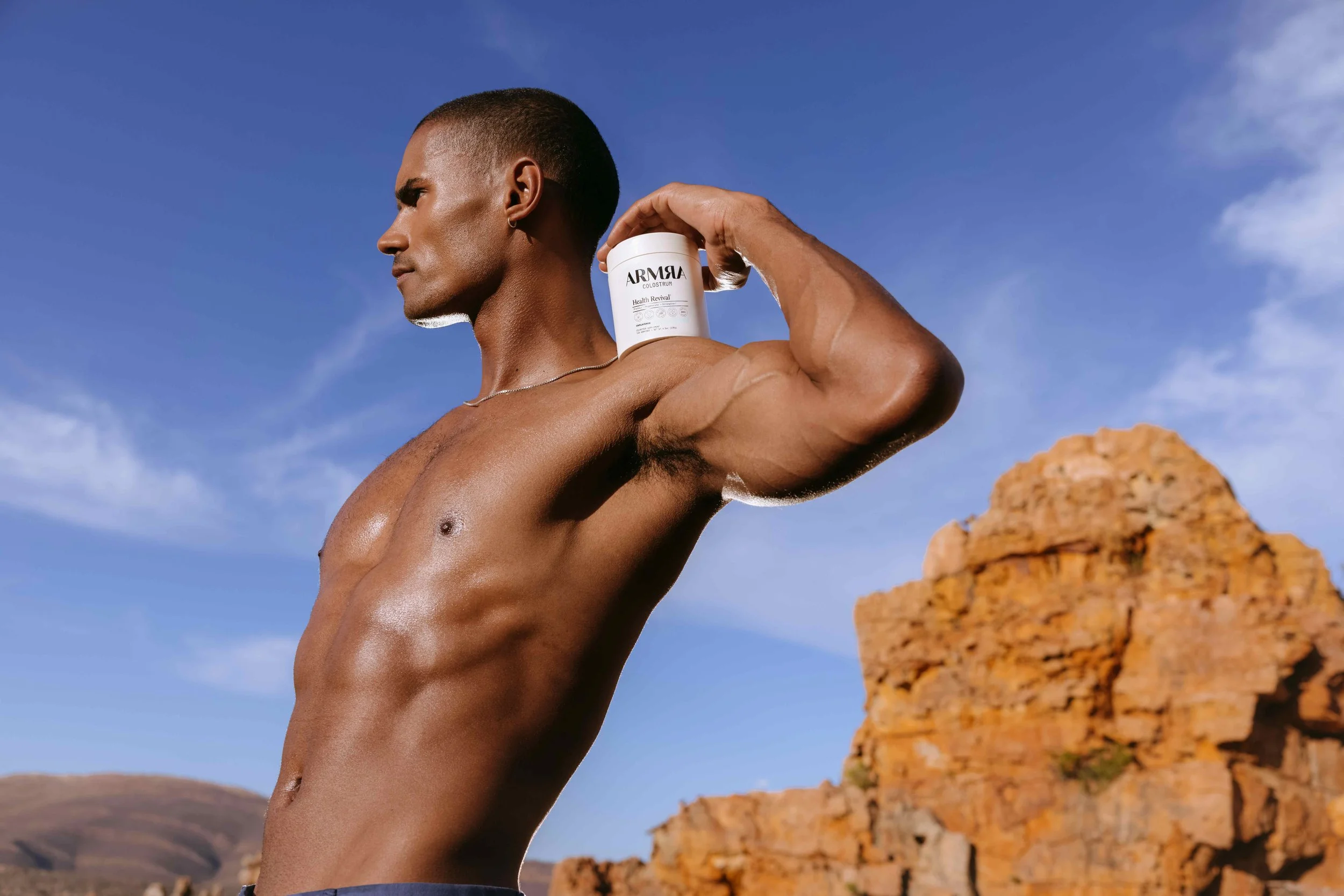 A shirtless man with short hair and a necklace holding a white bottle of ARMA Health Revival supplement outdoors against a blue sky and rocky landscape.