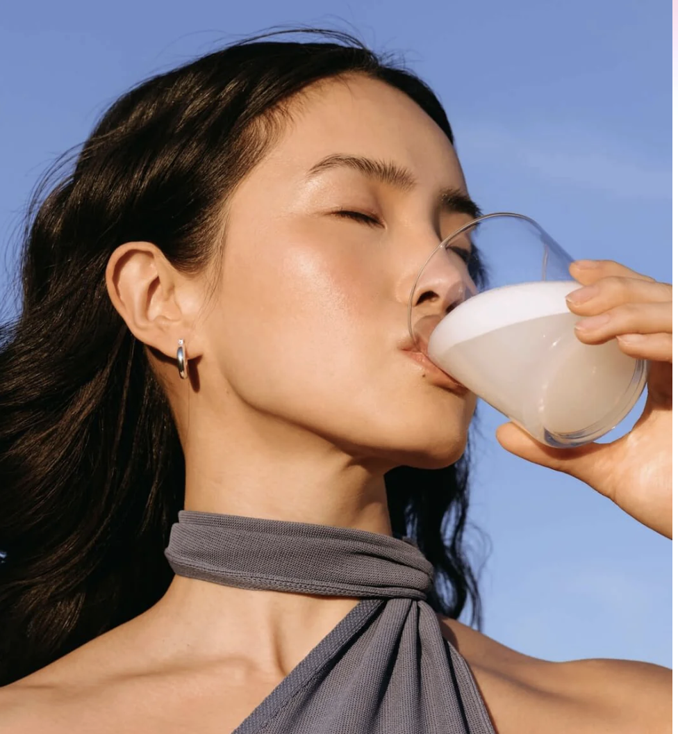 A woman with dark hair and hoop earrings drinks from a glass of milk against a clear blue sky.