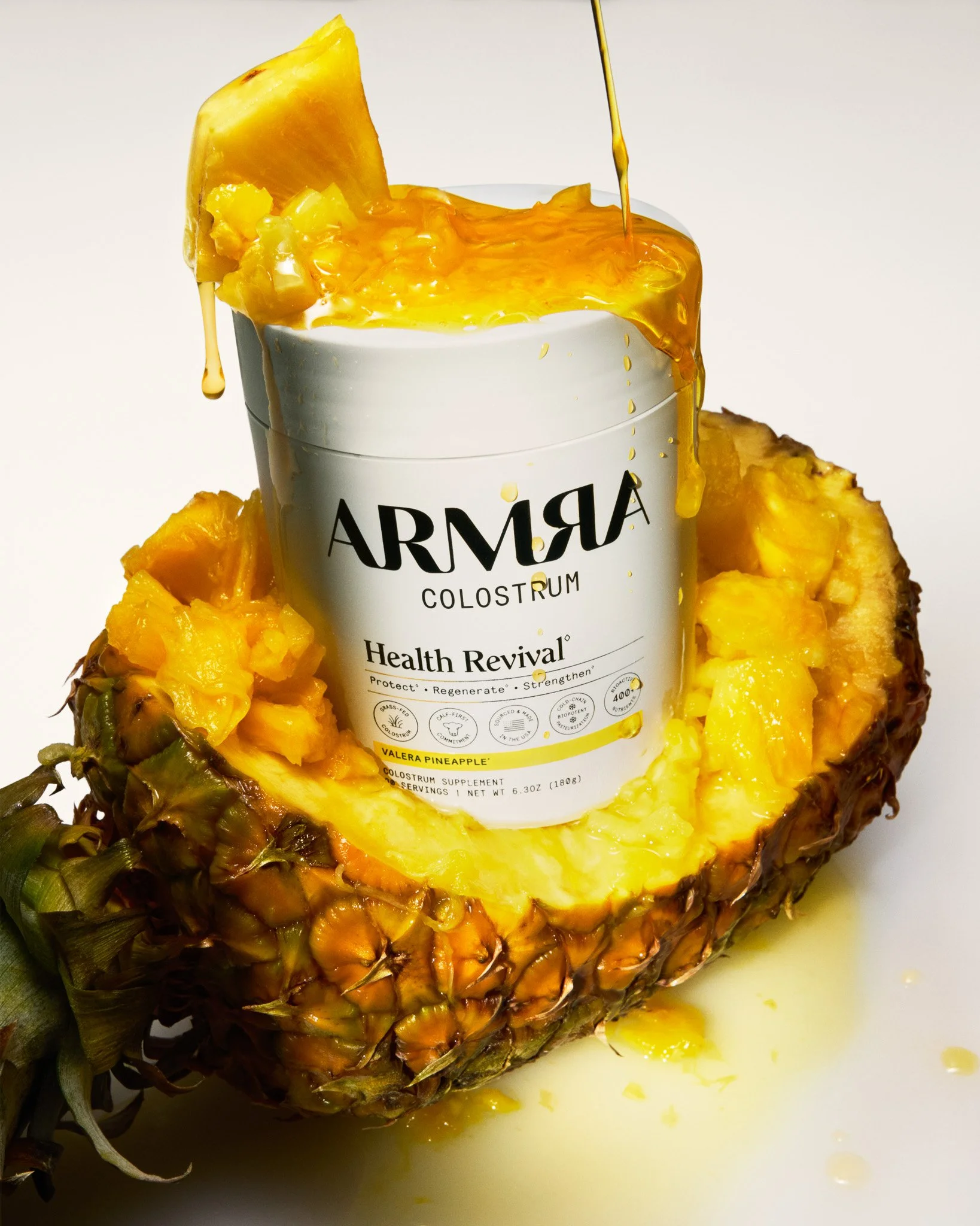 A pineapple with a container labeled 'Armra Colostrum' placed inside, with juice spilling over and pineapple pieces surrounding the container.