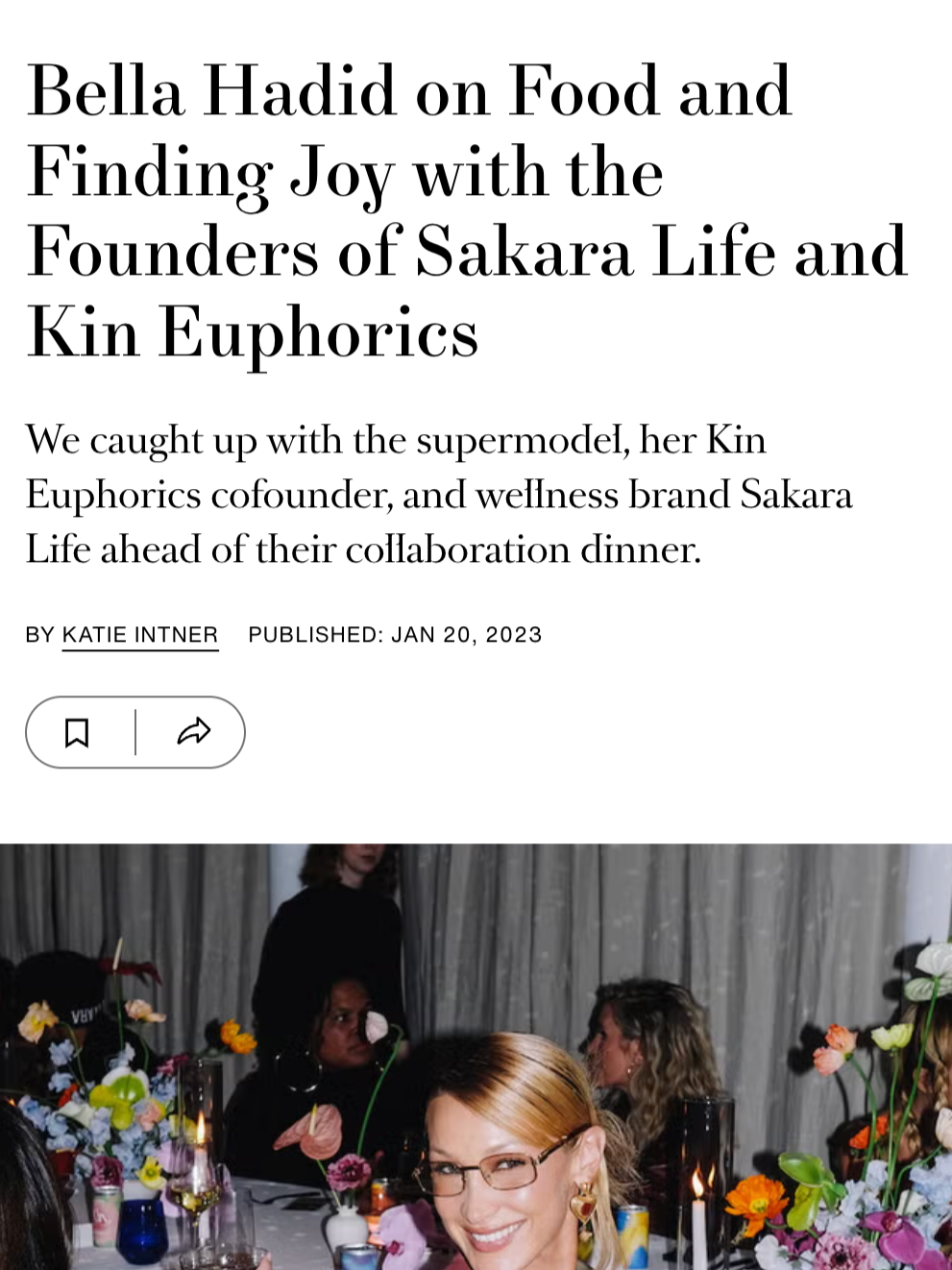A woman with red hair and glasses smiling at a dinner event with flowers and candles on the table, while other people are sitting around.