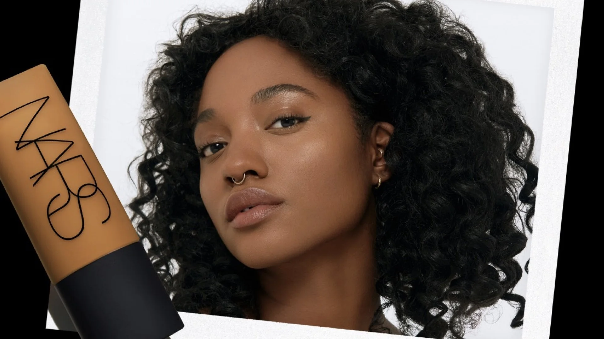 Close-up image of a woman with dark curly hair, gold hoop earrings, and a septum piercing, looking at the camera with a neutral expression, set against a white background with a large makeup product in the foreground. NARS