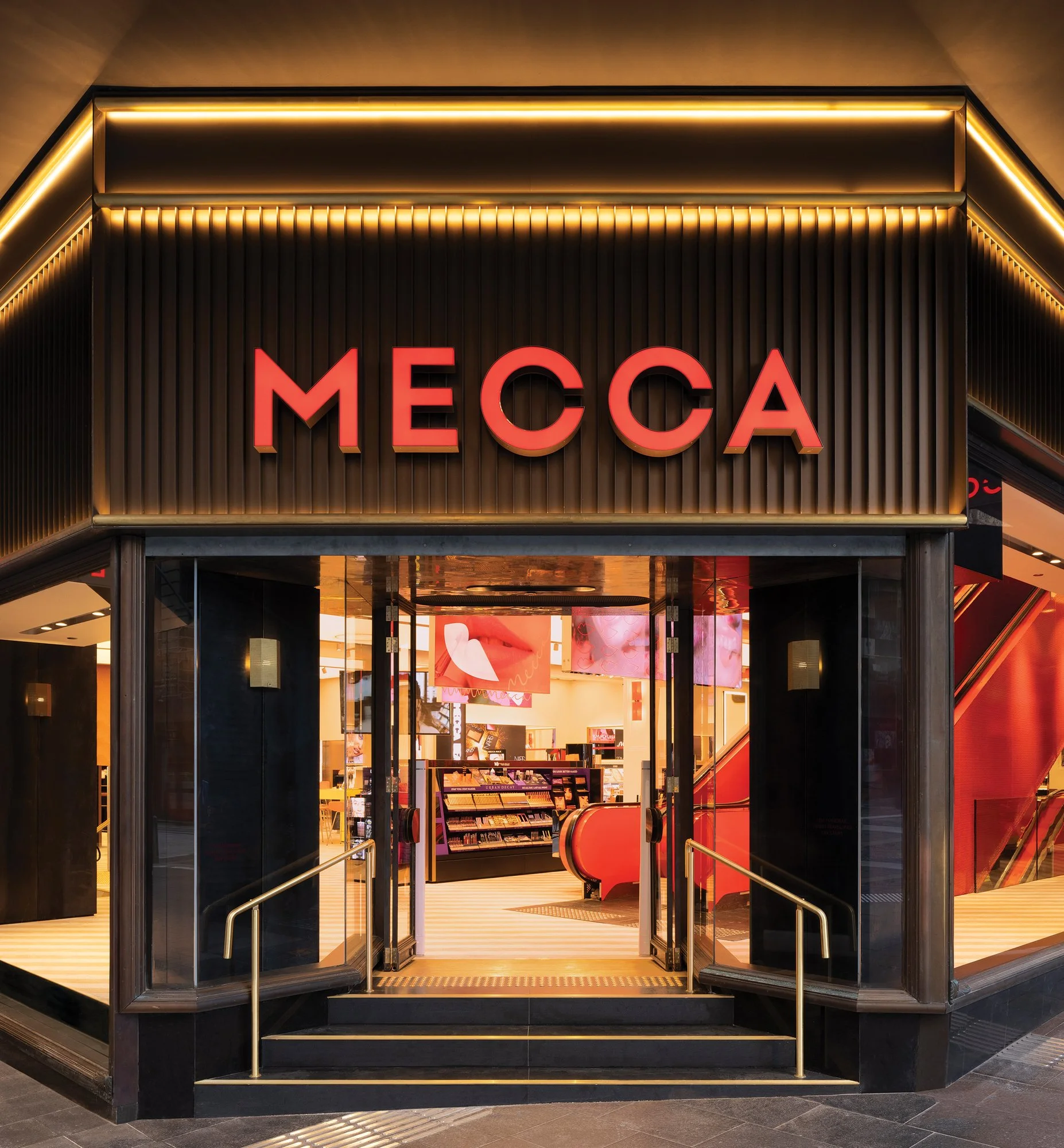 View of the entrance to MECCA, a cosmetics store in a shopping mall, with illuminated red sign and interior displays visible inside.