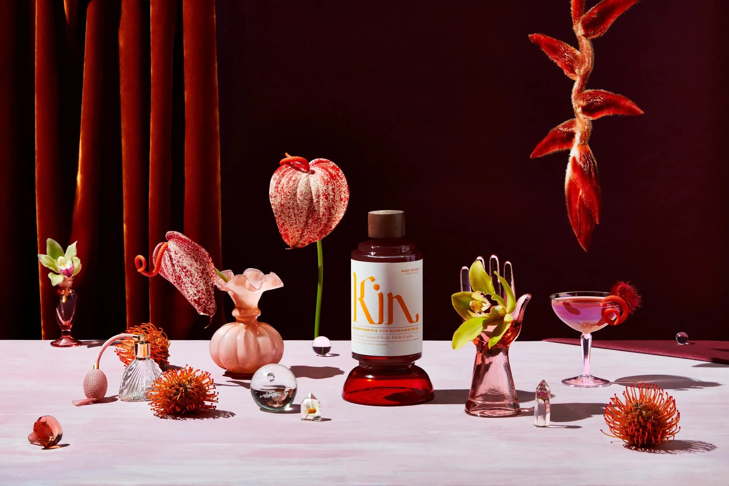 Arrangement of flowers, vases, glass balls, and a bottle with a label on a pink surface with a dark background.