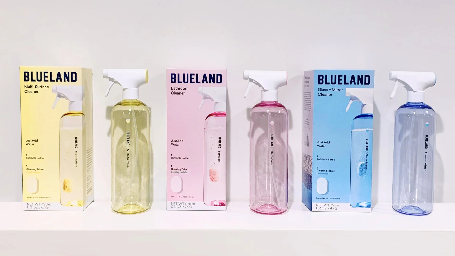 Four spray bottles with their boxes behind them, containing different cleaning solutions in various colors, on a white surface with a plain white wall background.