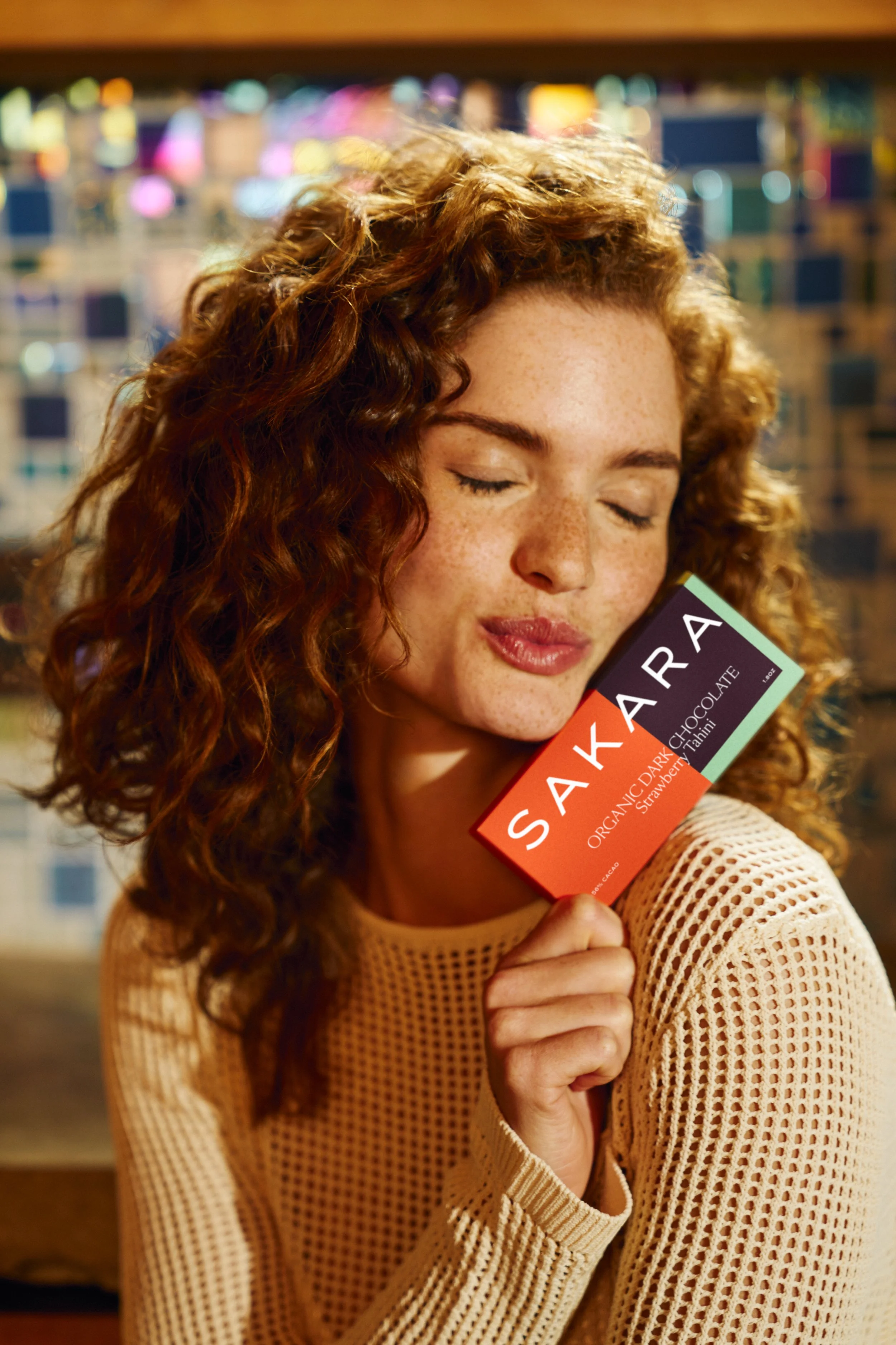 A woman with curly red hair and freckles is holding a Sakara chocolate bar close to her cheek with her eyes closed.