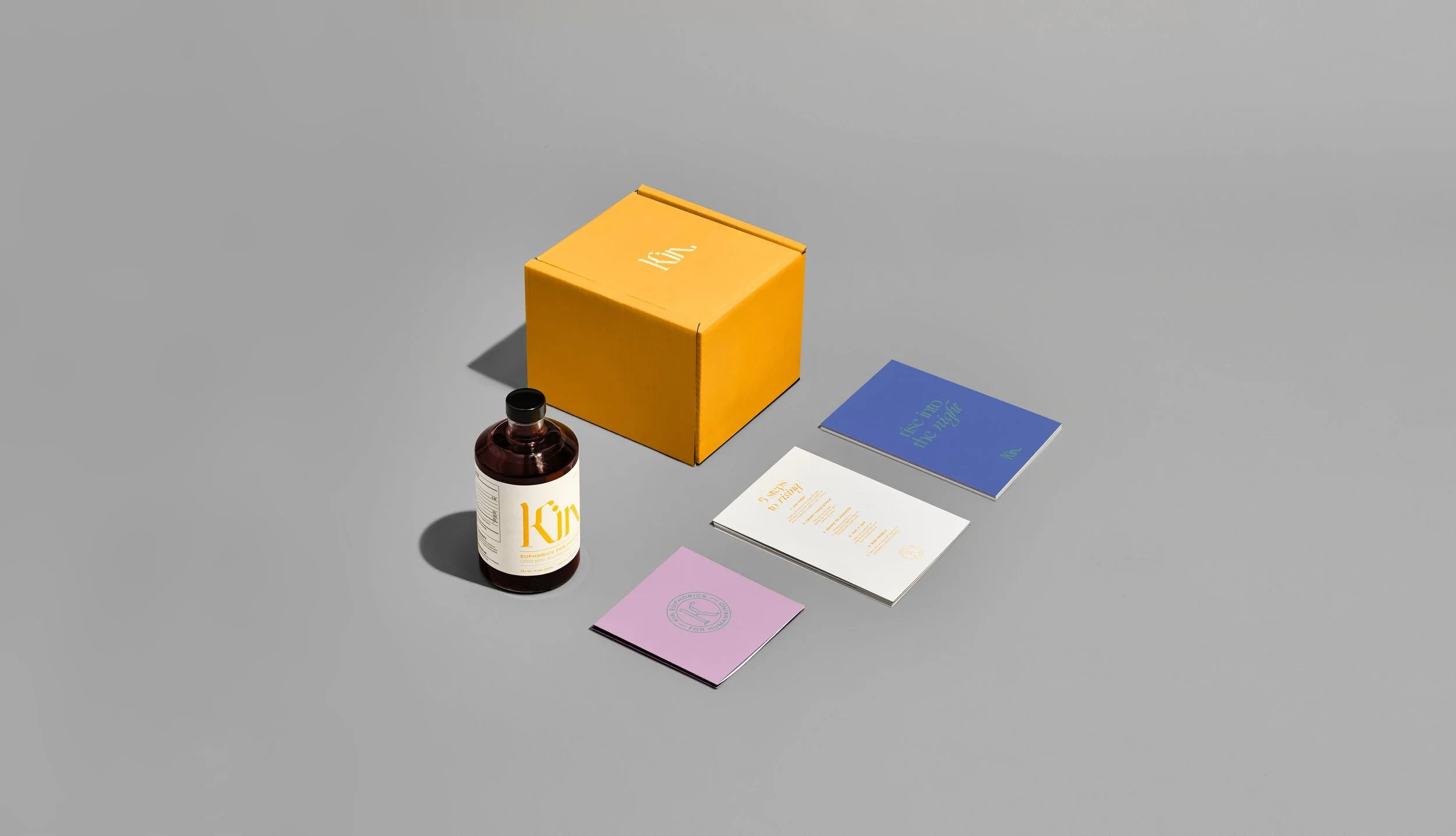 A yellow boxed product, a brown bottle labeled 'Kym', and three small booklets in different colors (purple, white, and blue) on a gray surface.