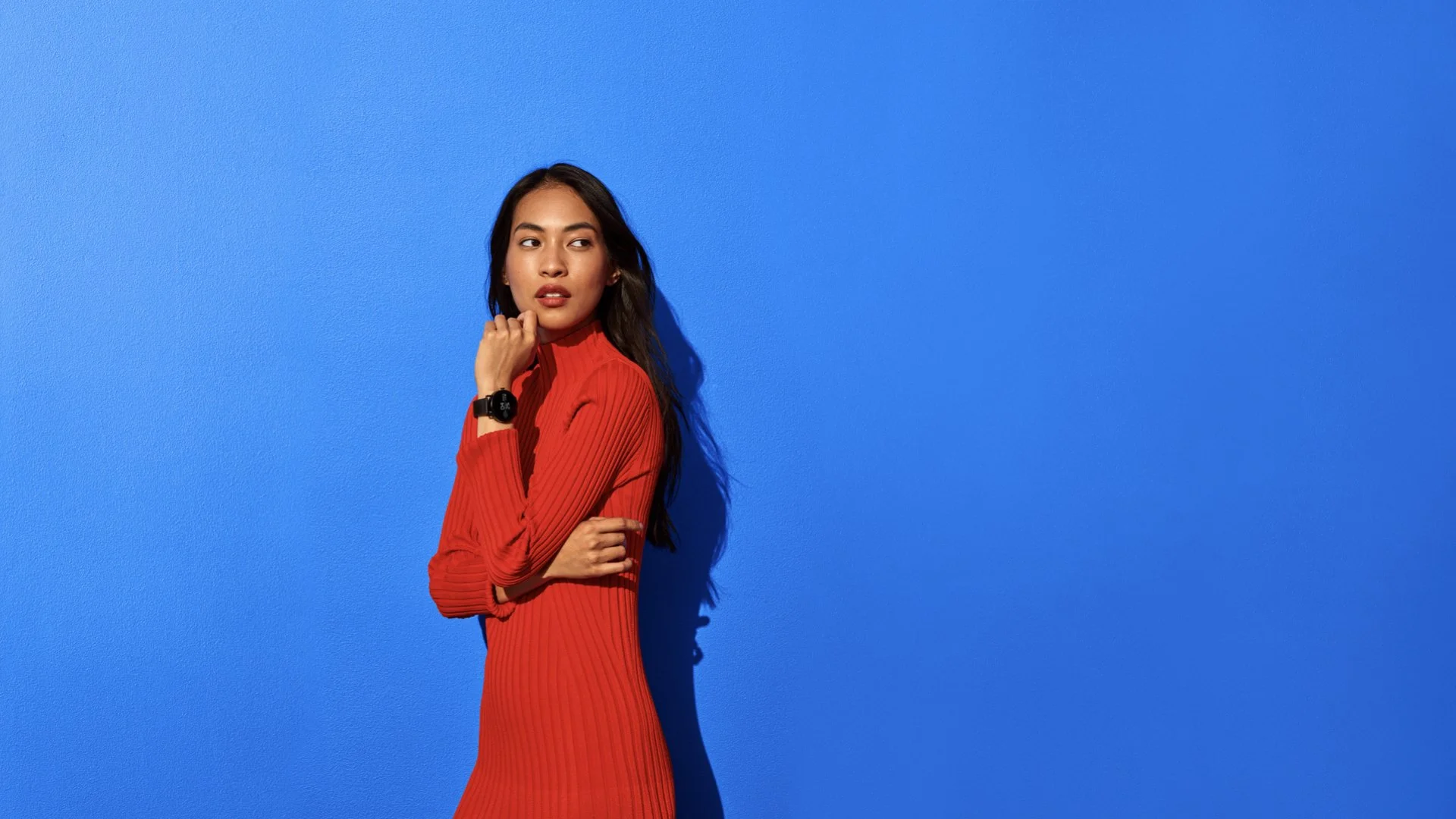 A woman with long dark hair wearing a red dress and black watch standing against a bright blue wall.