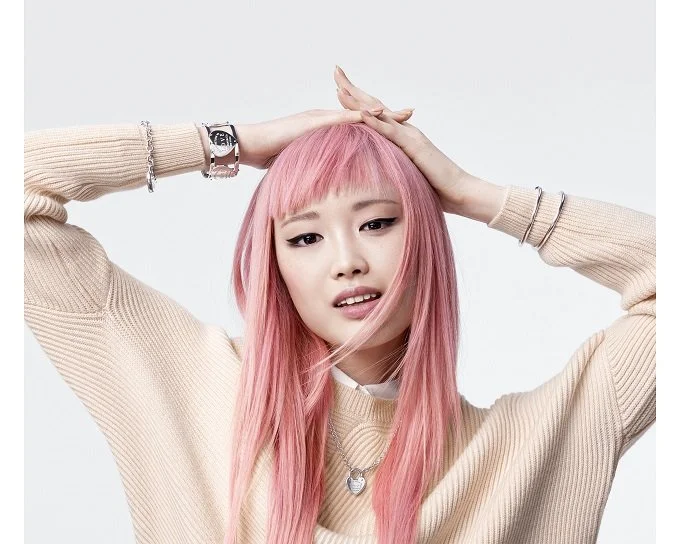 Young woman with pink hair in a beige sweater holding her head with one hand and looking confused or worried.