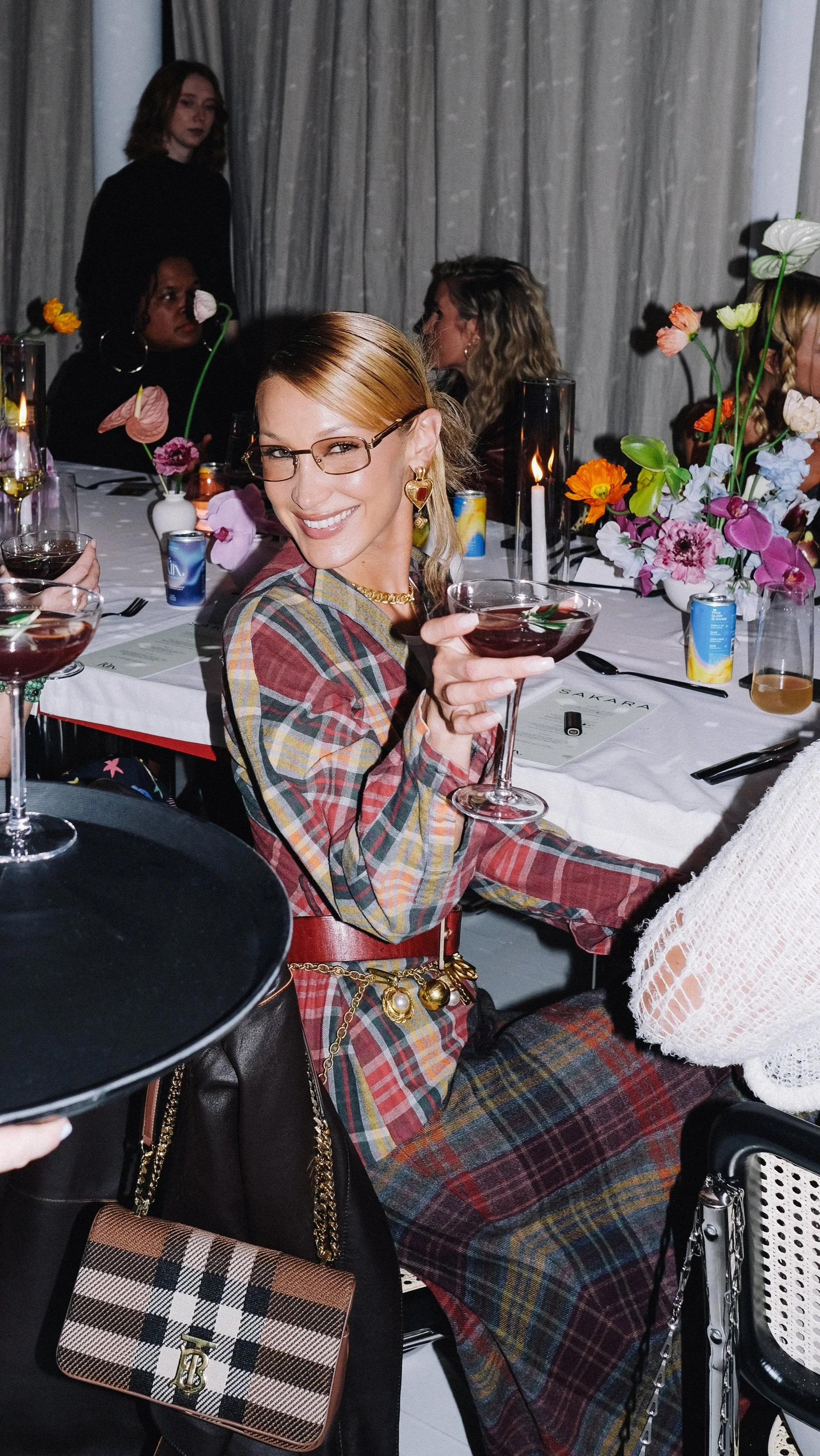A woman wearing glasses and gold jewelry sitting at a table, smiling and holding a glass of red wine in a party or celebration setting with other women and floral arrangements.
