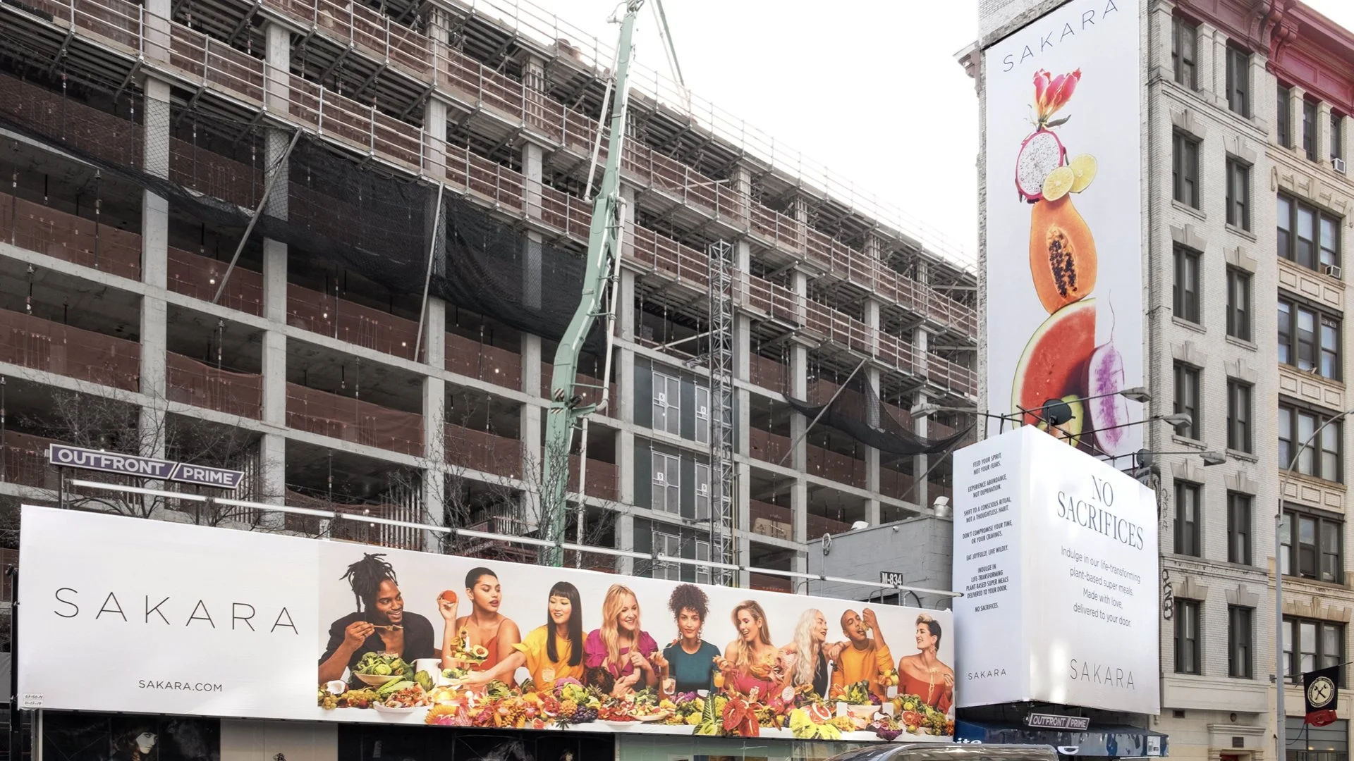 Large billboard advertisements on building construction site featuring diverse group of women eating and enjoying food, promoting SAKARA brand and plant-based meals.
