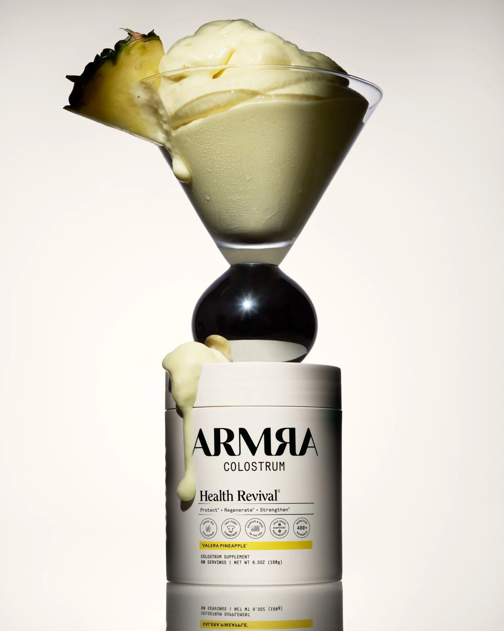 A container labeled 'ARMRA Colostrum Health Revival' with a scoop of colostrum powder on top, a glass of whipped creamy substance garnished with a piece of pineapple, a black and silver ball, and a droplet of white substance dripping from the contain