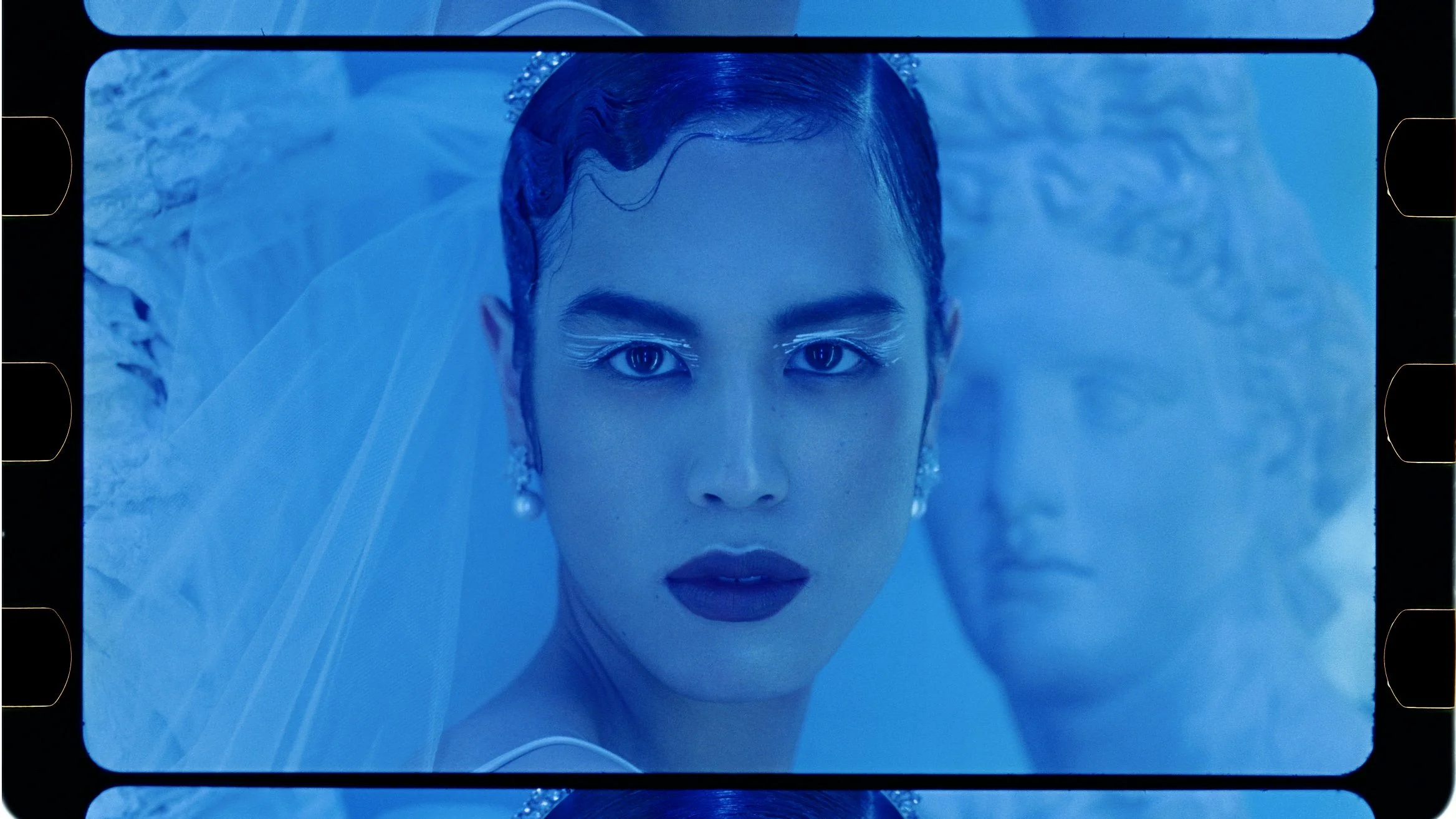 Close-up of a woman with dramatic makeup, earrings, and a veil, looking directly at the camera with a man behind her, both in a blue-tinted setting.