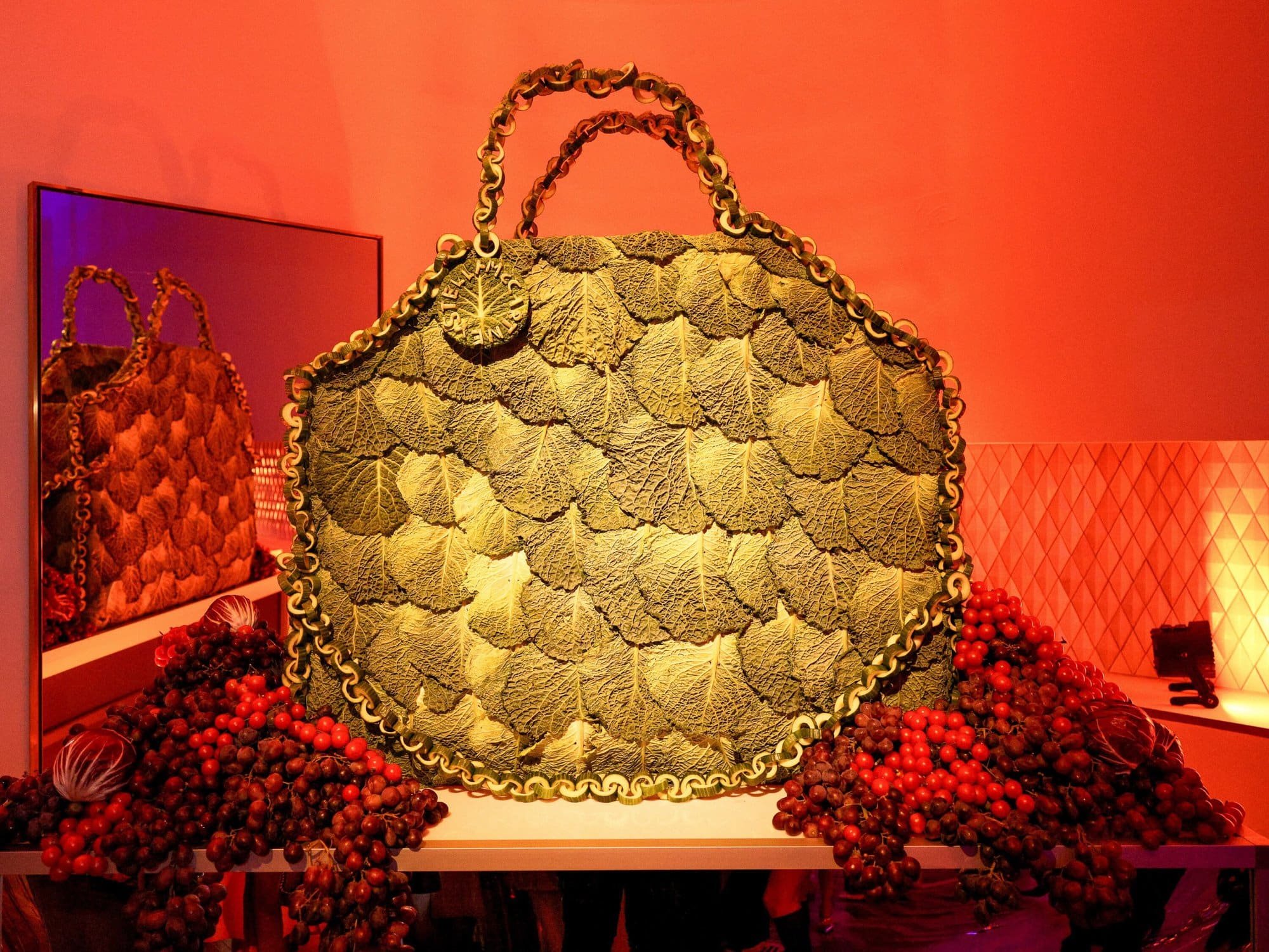 A large handbag covered with green leaves and surrounded by red and purple berries, displayed on a table at an exhibition with red lighting.