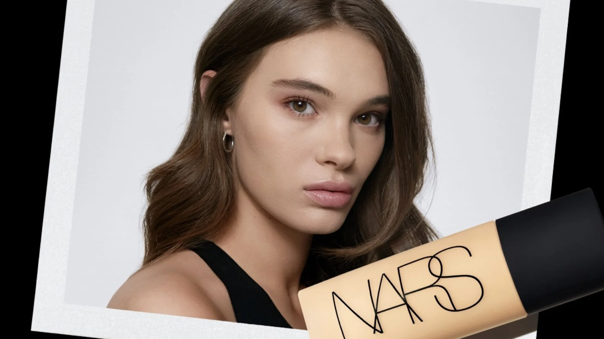 A young woman with brown hair and light makeup, gazing at the camera, holding a NARS foundation bottle.