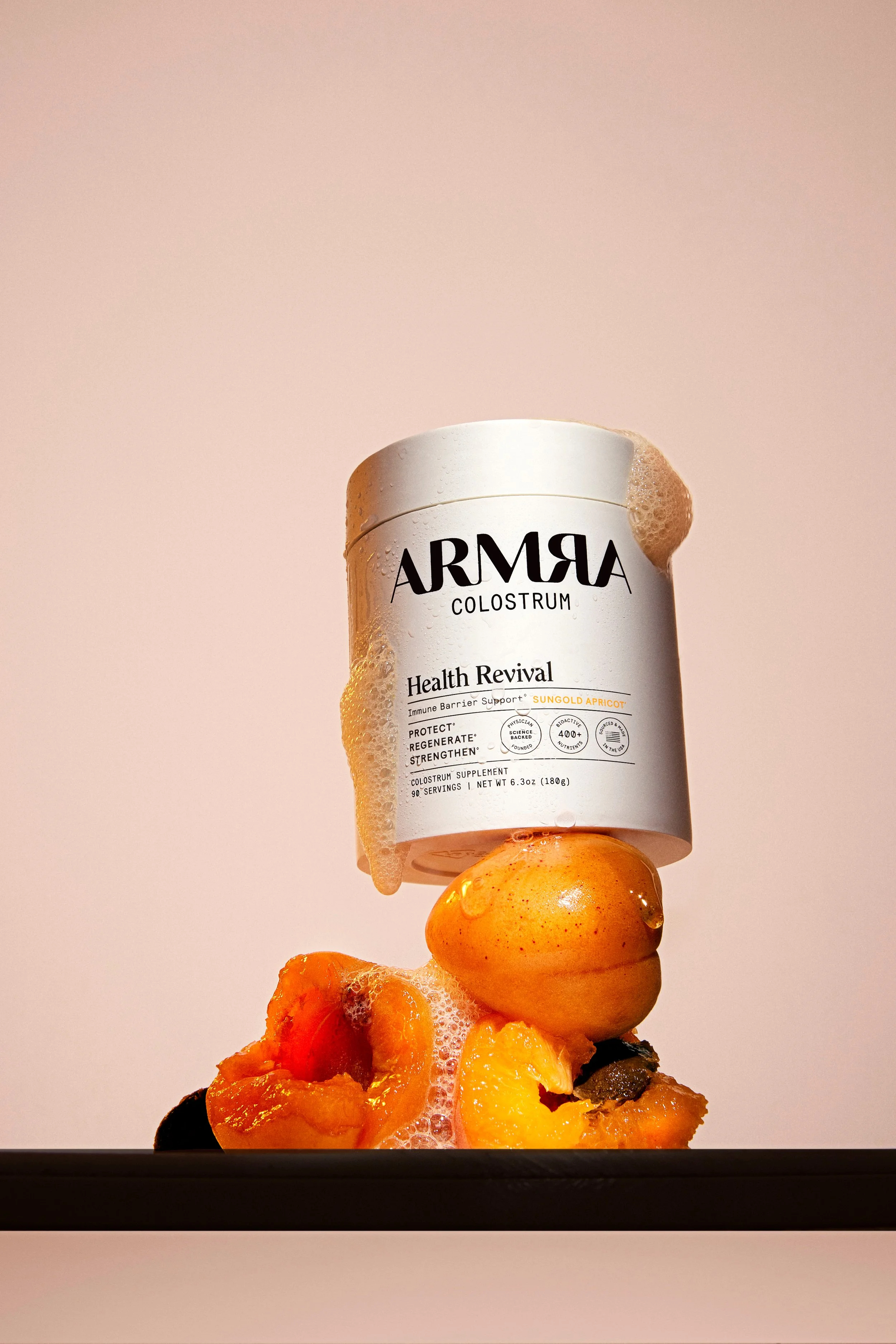 A container of ARMA Colostrum Health Revival placed balanced on a small fruit with foam bubbles and soap suds on the fruit and container, against a plain light pink background.
