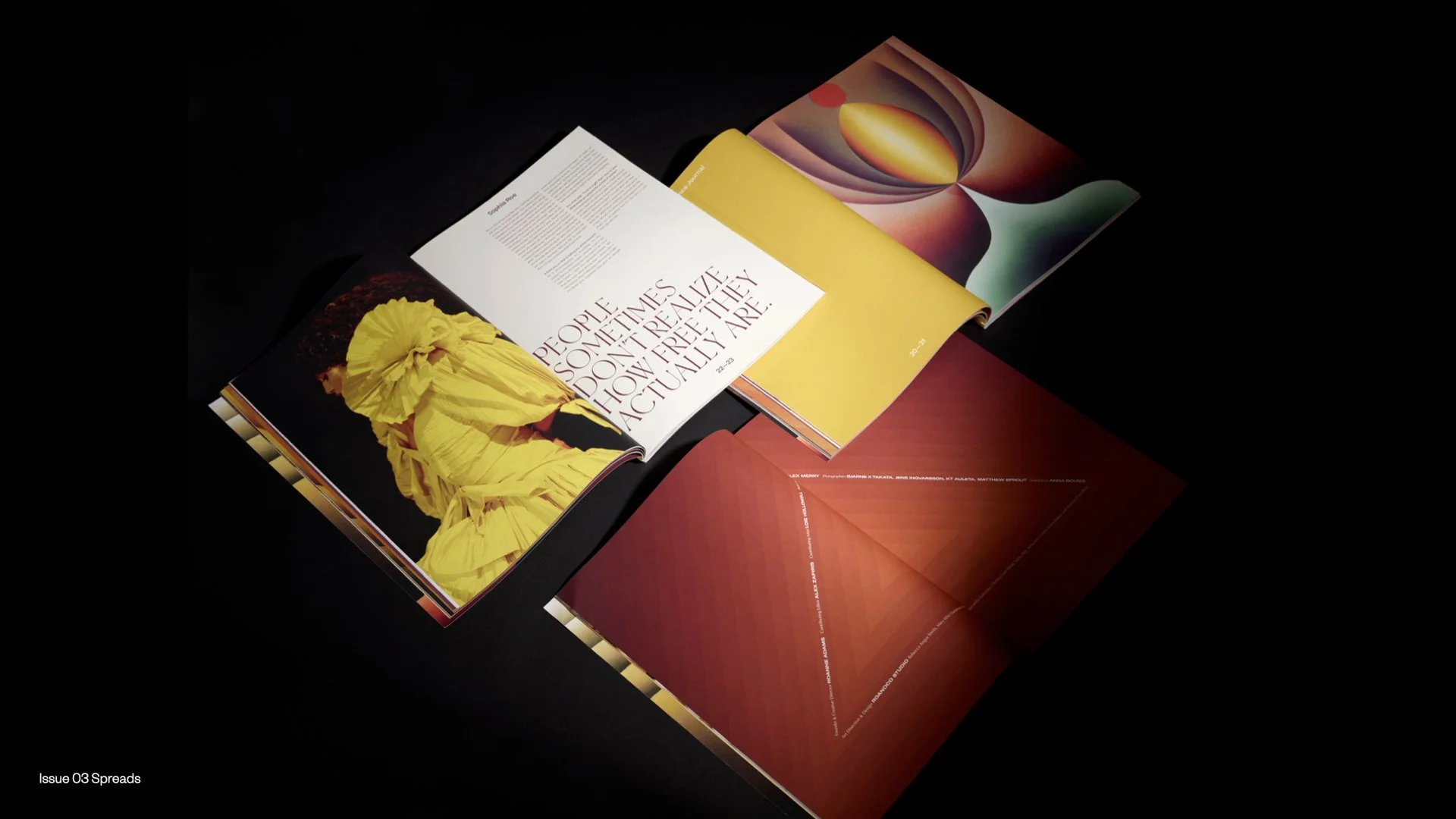 Open magazines spread out on a black surface, featuring colorful abstract art, a photograph of a person in a yellow outfit, and large text saying "People sometimes don’t realize how freely they actually are."