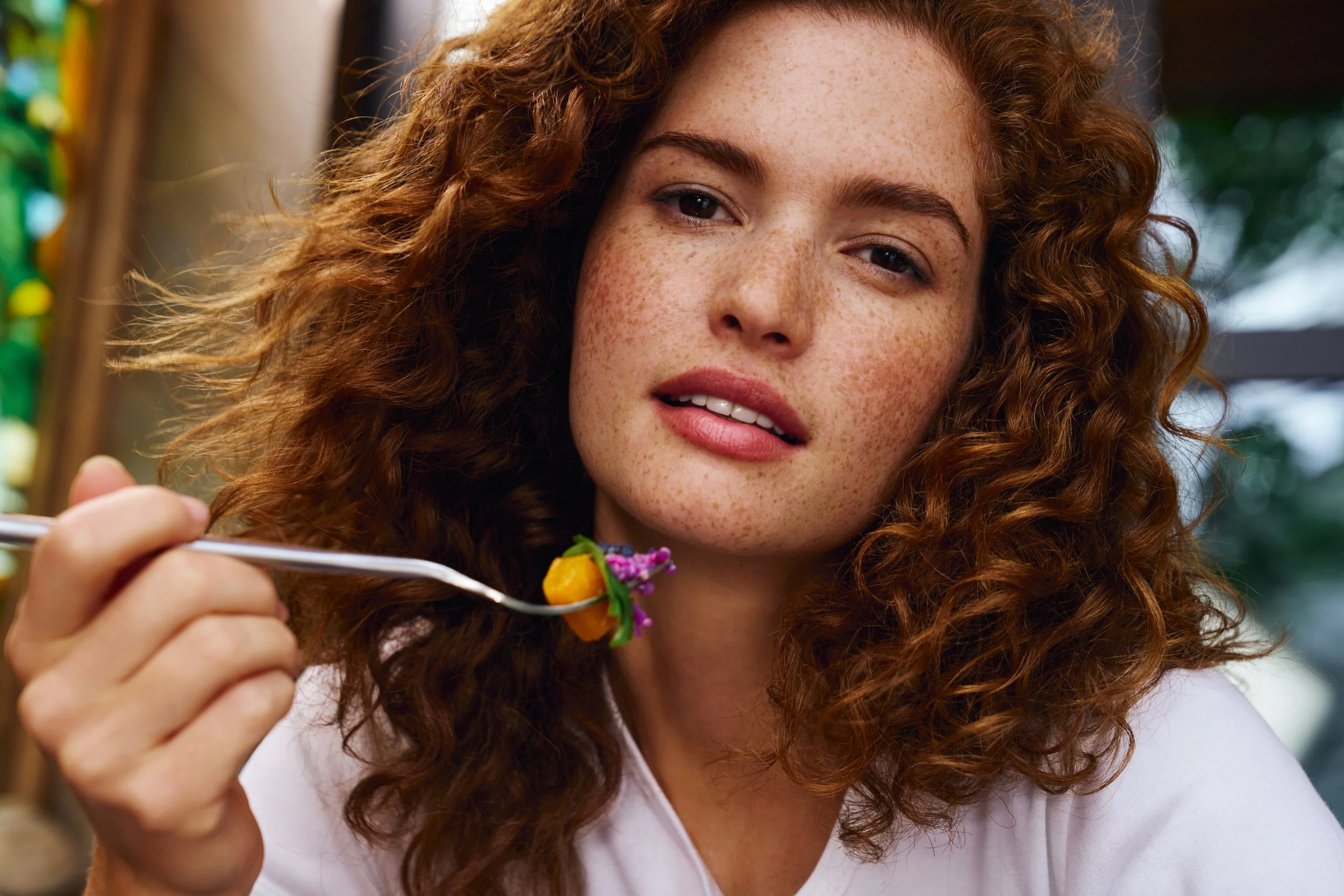 A young woman with curly red hair and freckles is holding a fork with food close to her mouth, looking at the camera, in a brightly lit indoor setting.