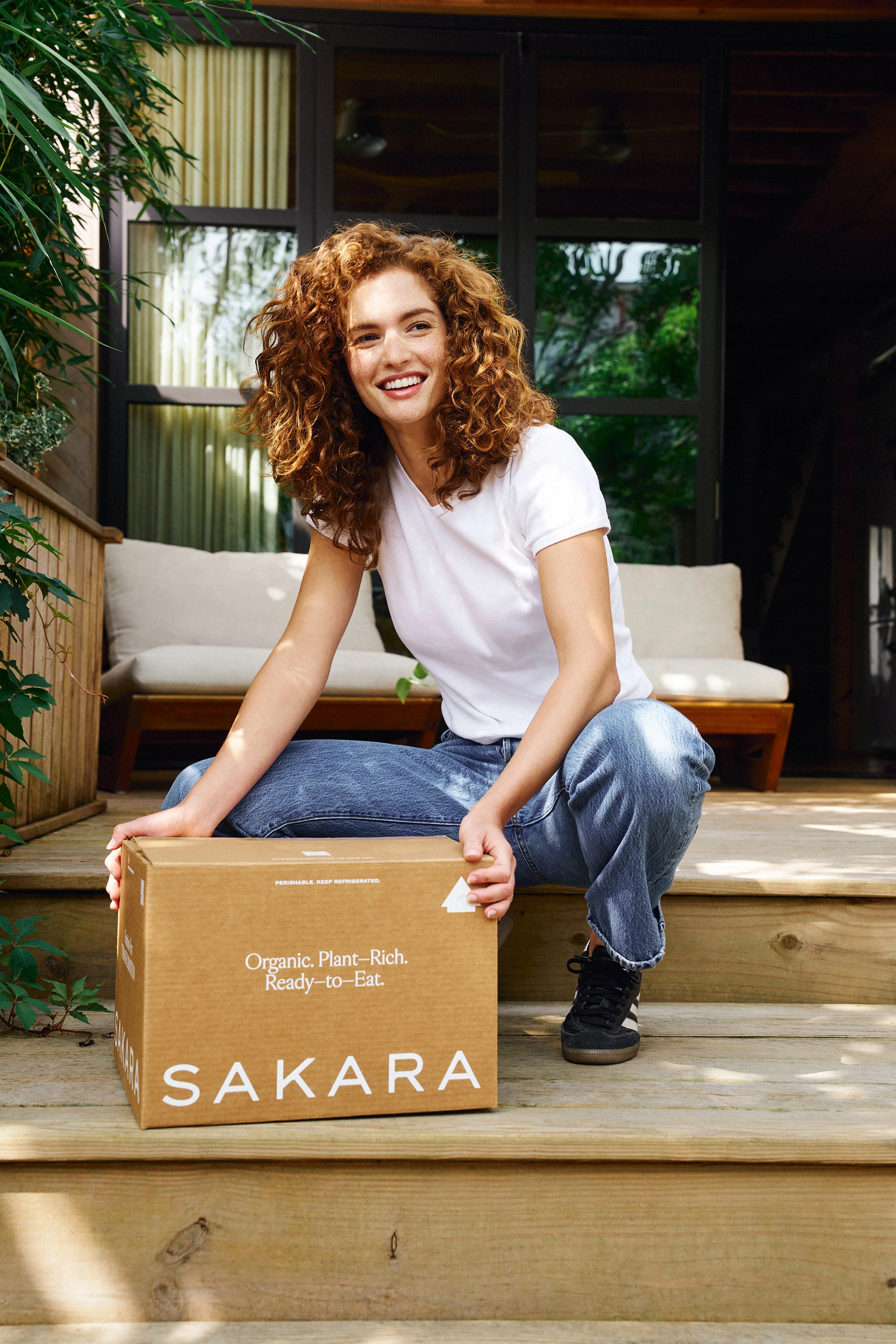 A woman with curly red hair, wearing a white t-shirt and jeans, sitting on wooden stairs outside a house. She is smiling and holding a cardboard box labeled "SAKARA," which contains organic, plant-rich, ready-to-eat food.