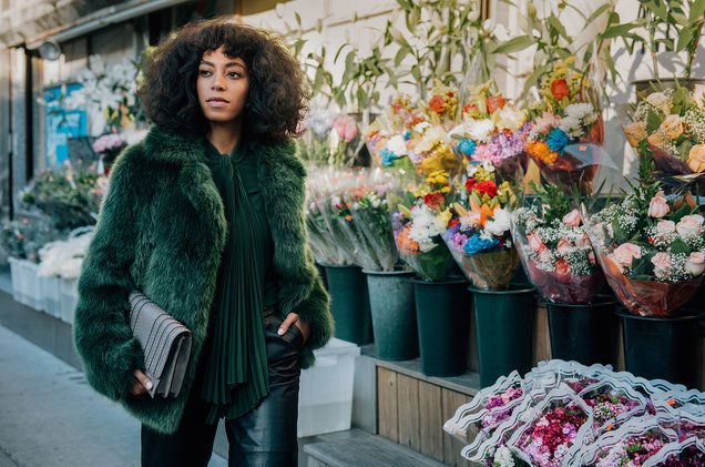 A woman with curly hair in a green fur coat stands in front of a flower shop, holding a clutch purse. Solange Knowles for Michael Kors