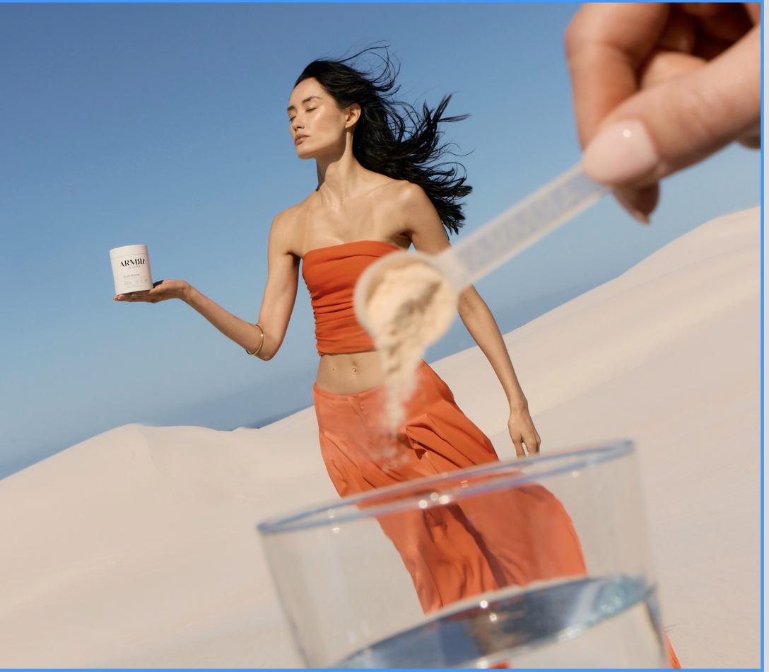 A woman in a strapless orange top and matching skirt stands on sand dunes with a clear blue sky, holding a container of skincare product, while a hand in the foreground adds a scoop of powder into a glass of water.